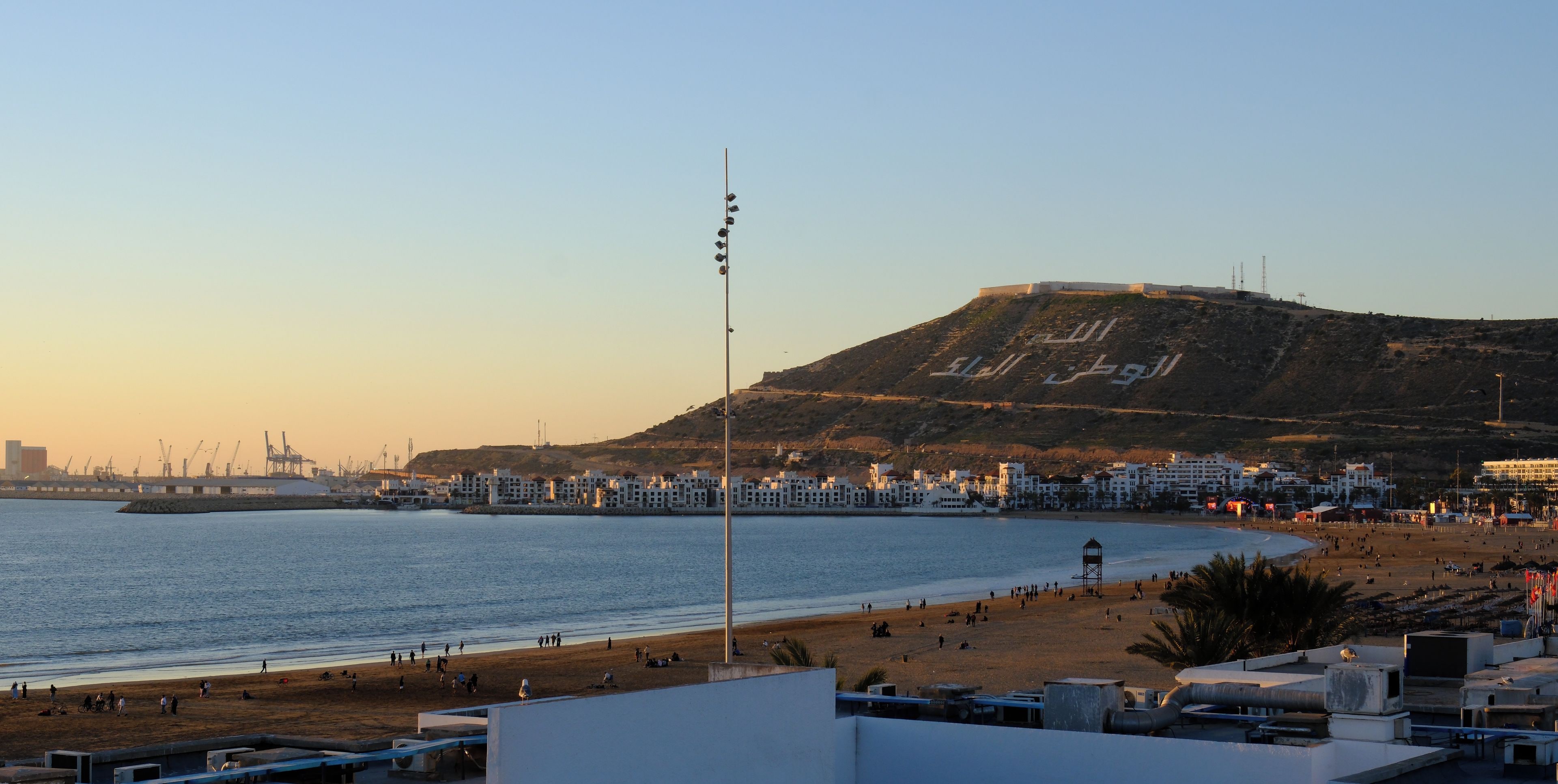 A coastal cityscape with a beach, a hill with inscriptions, and a clear sky.
