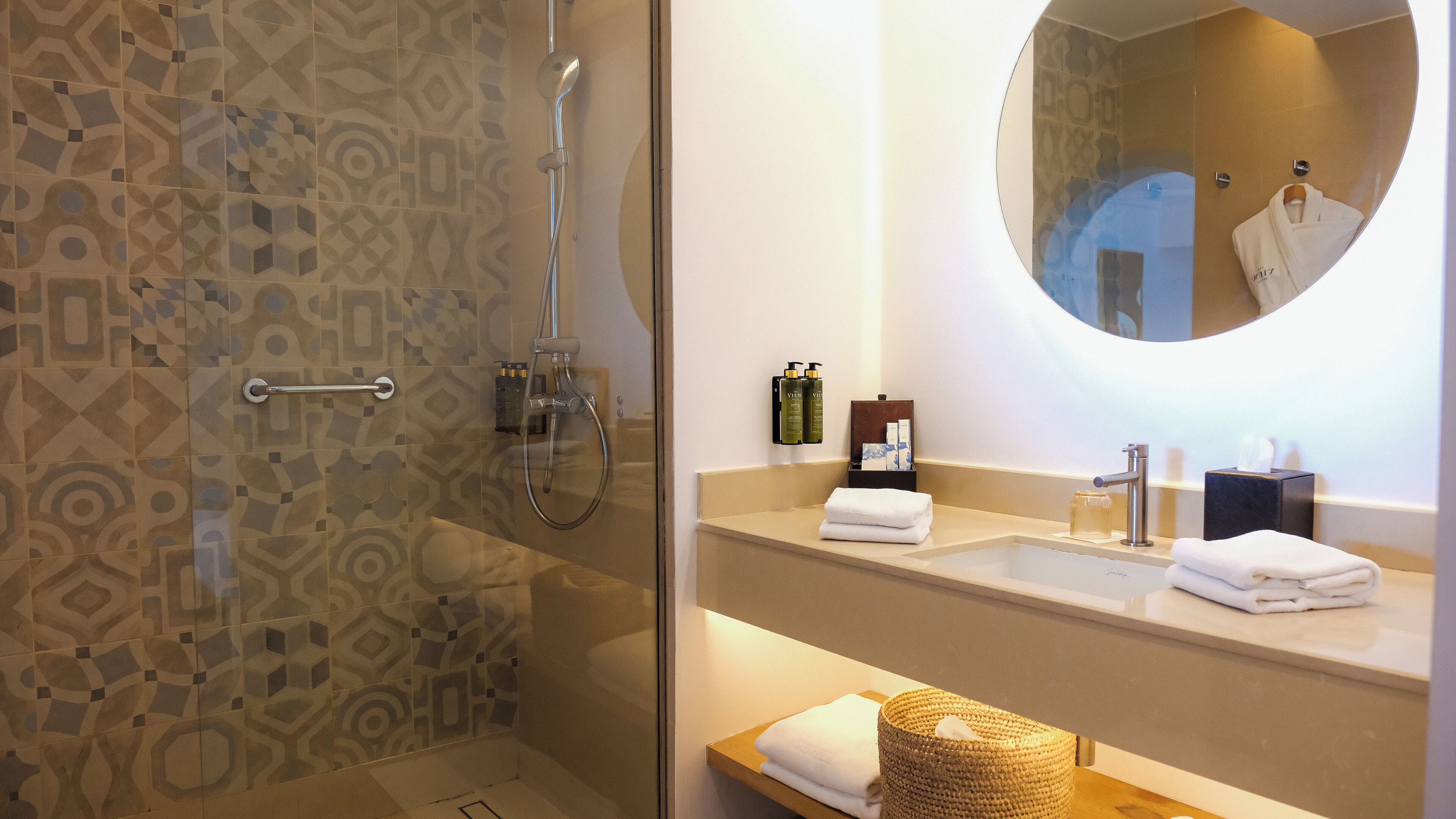 Un baño moderno con baldosas estampadas, ducha con mampara de vidrio, espejo redondo y tocador con lavamanos, toalla y artículos de aseo.