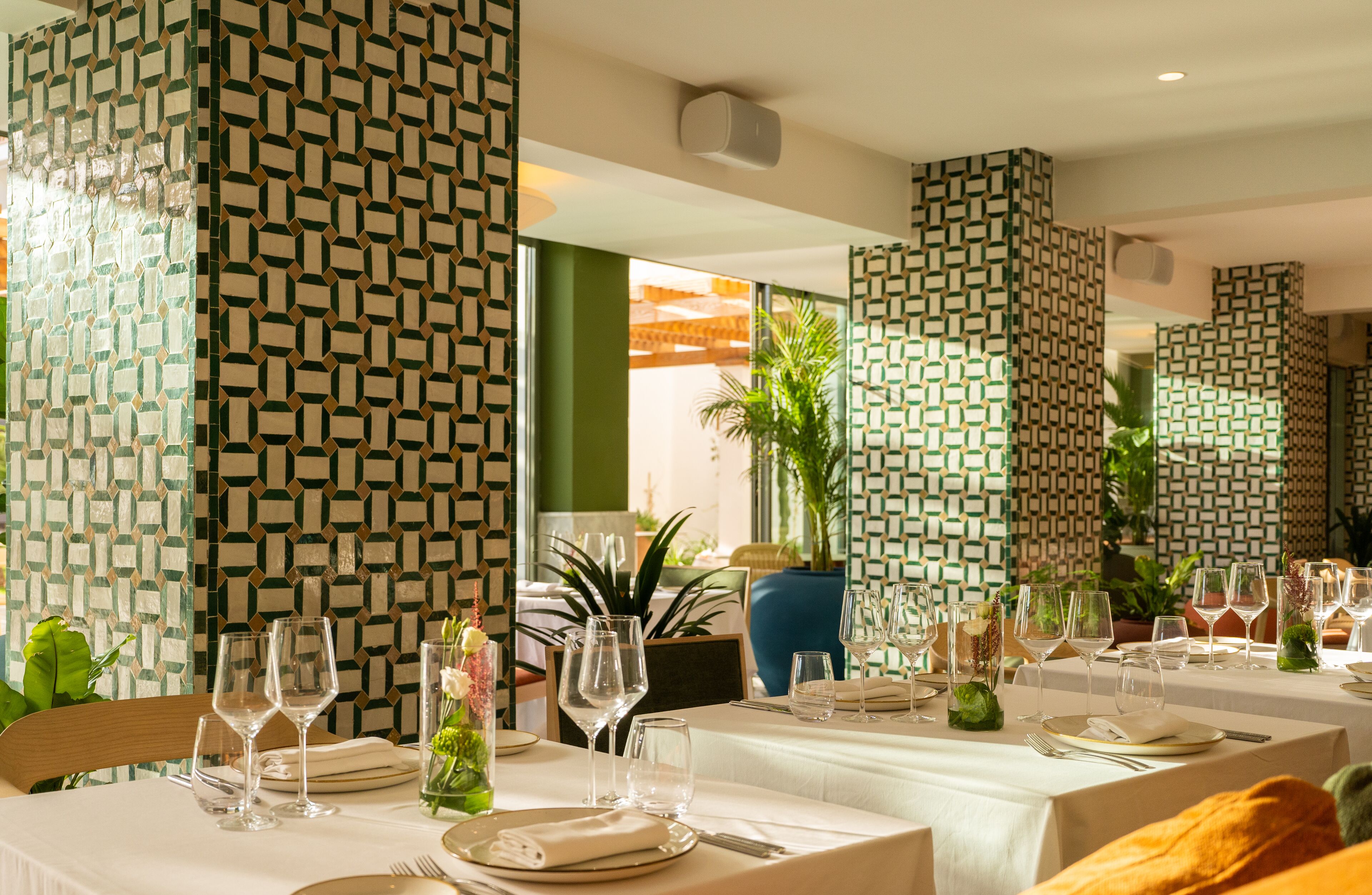 A restaurant with a geometric patterned wall, tables set with white tablecloths, and empty chairs.