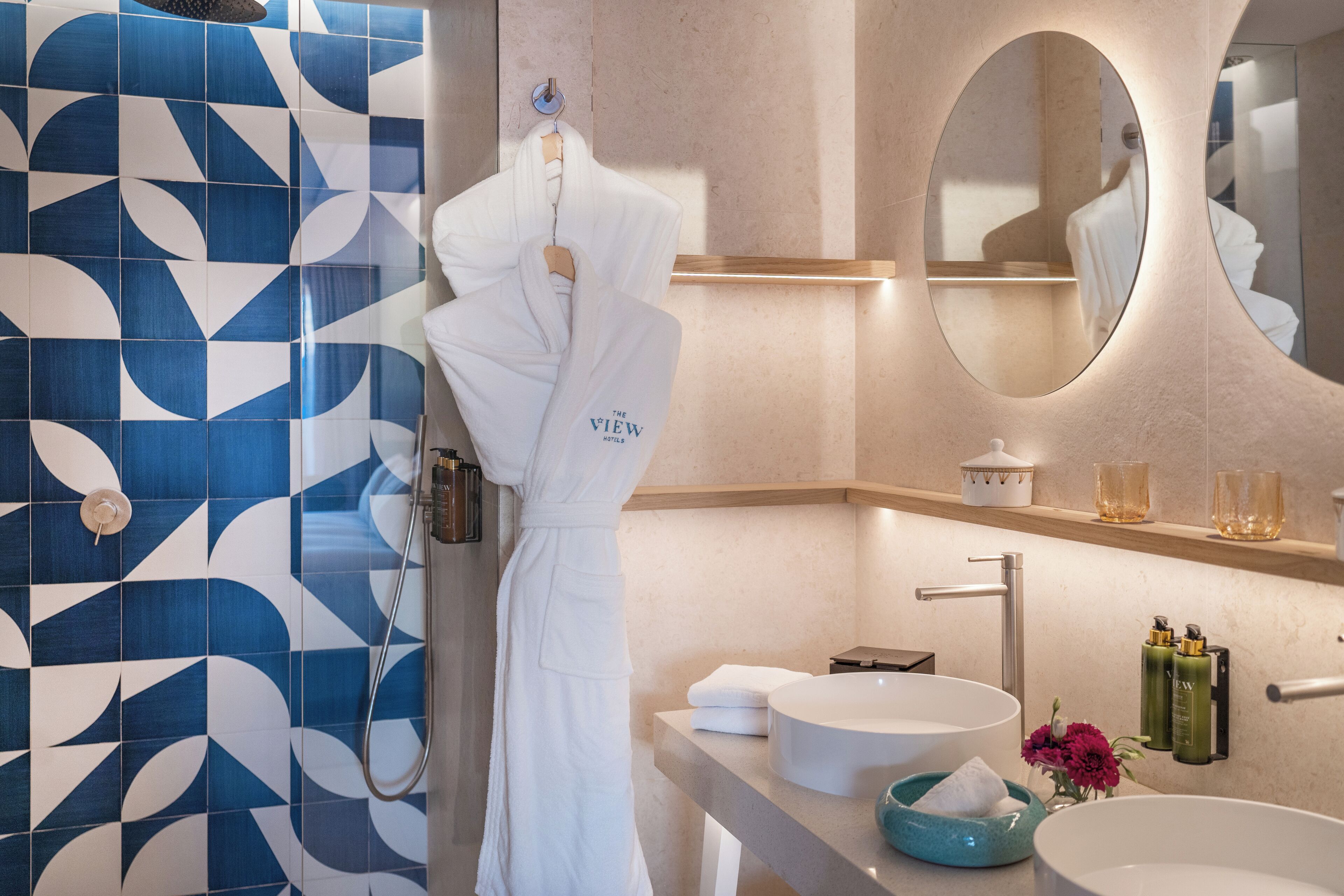 Une salle de bain moderne avec un carrelage bleu et blanc, un grand miroir, un lavabo avec un récipient bleu et un peignoir suspendu à un porte-serviettes.
