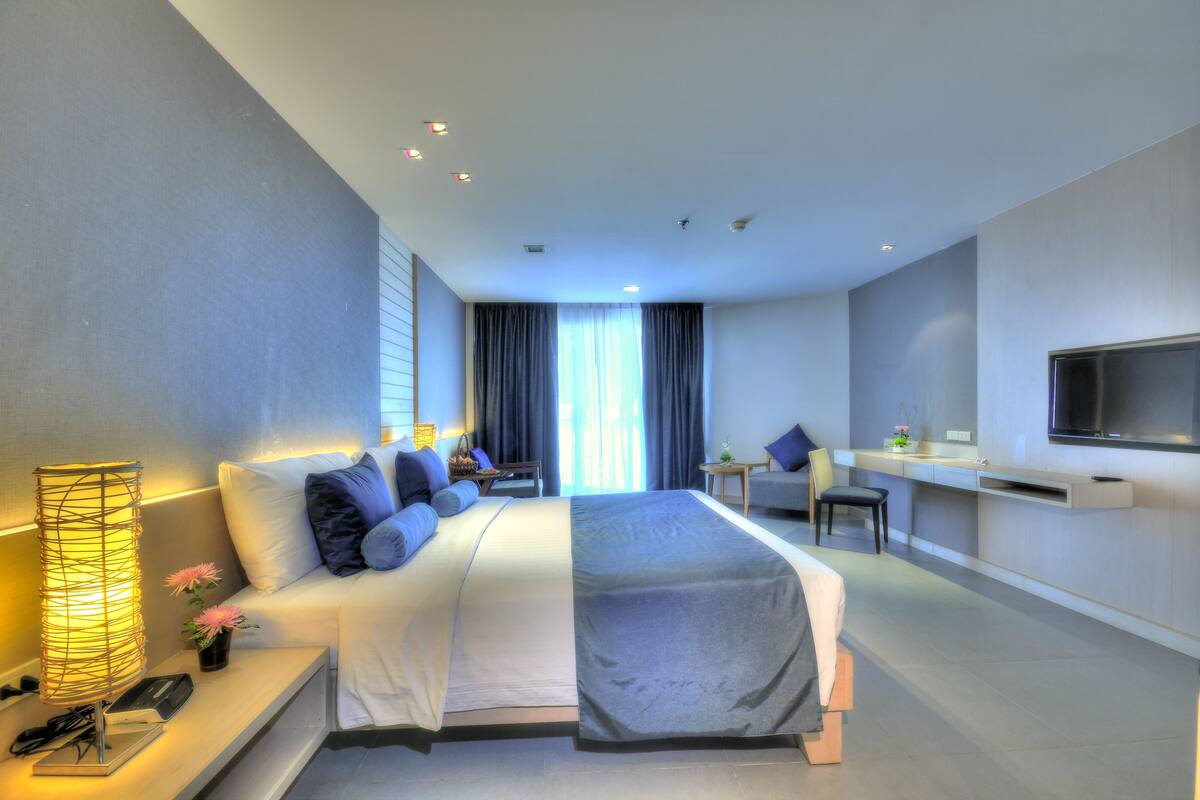 A modern hotel room with a large bed, bedside table, lamp, TV, and a desk area.