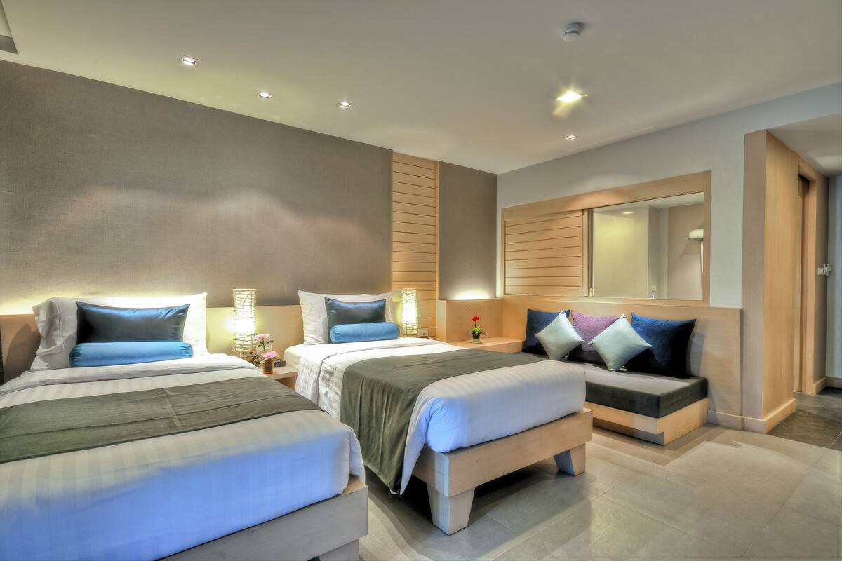 A modern hotel room with three beds, a large mirror, and a neutral color scheme.