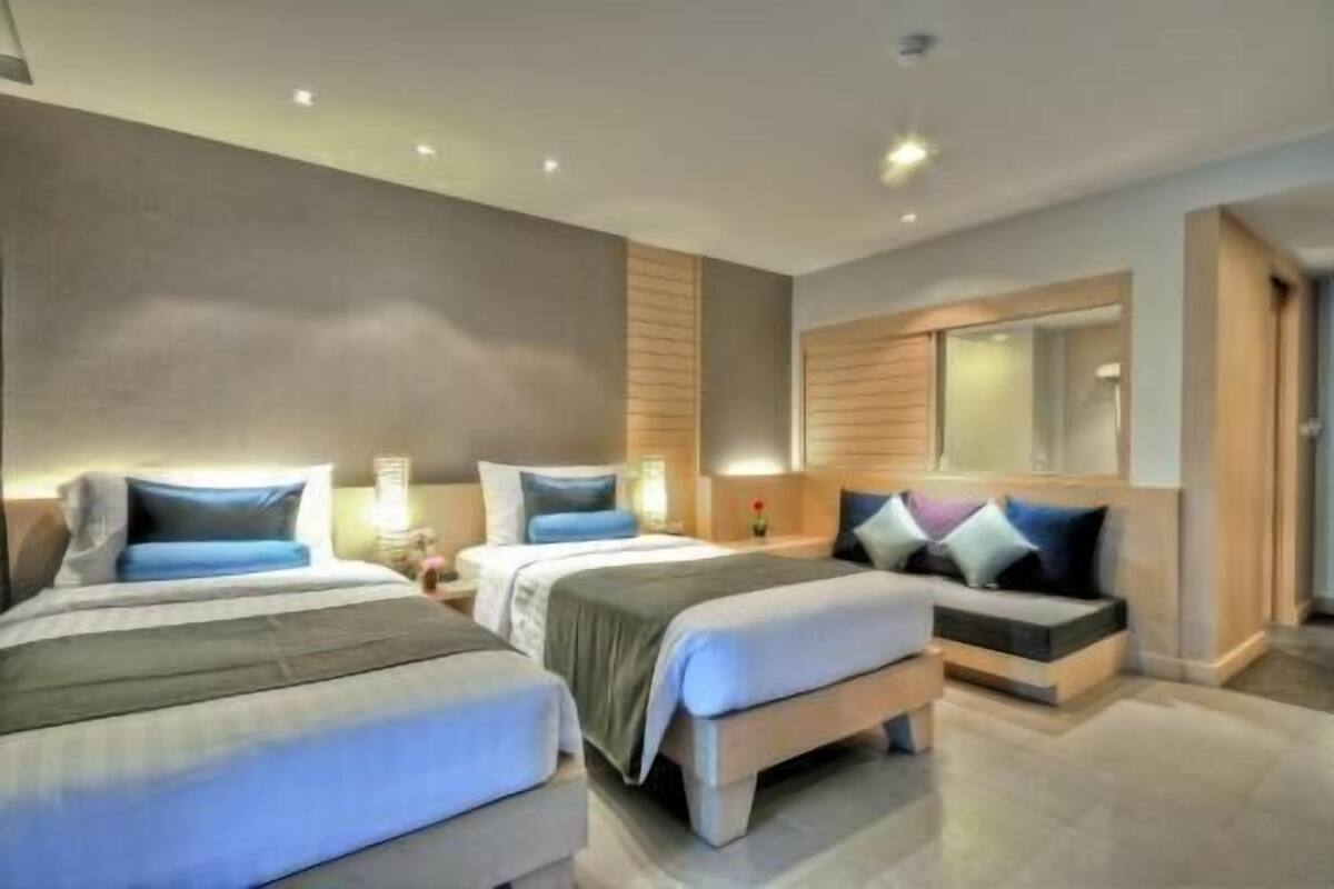 A modern hotel room with three beds, a large mirror, and a neutral color scheme.