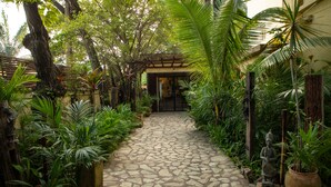 A stone pathway leading to a building surrounded by lush tropical plants and a statue.