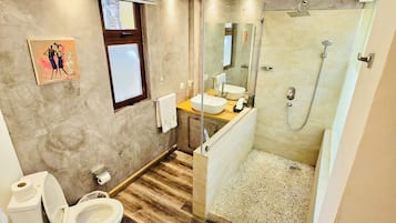 Un baño moderno con suelo de madera, un inodoro, un lavabo, una ducha con puerta de cristal y una ventana pequeña.