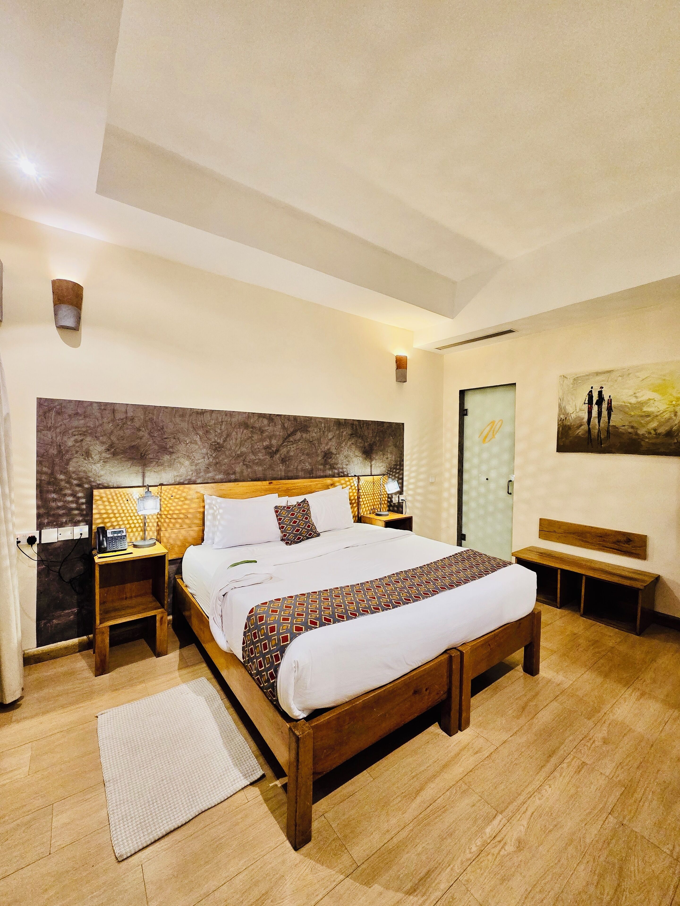 A modern hotel room with a large bed, wooden headboard, bedside tables with lamps, a bench, and a painting on the wall.