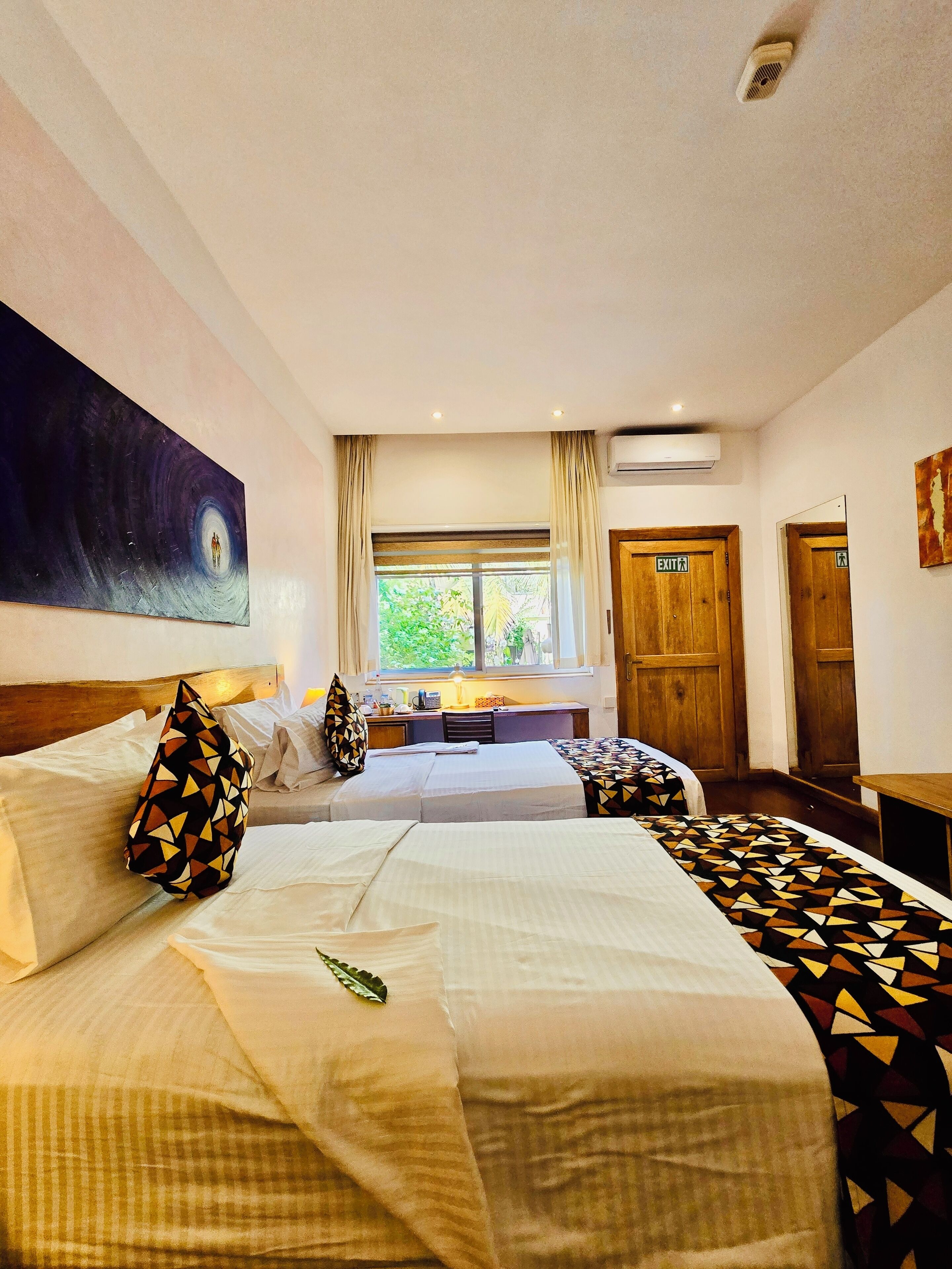 A hotel room with two beds, a large painting on the wall, a desk with a lamp, and a window with curtains.