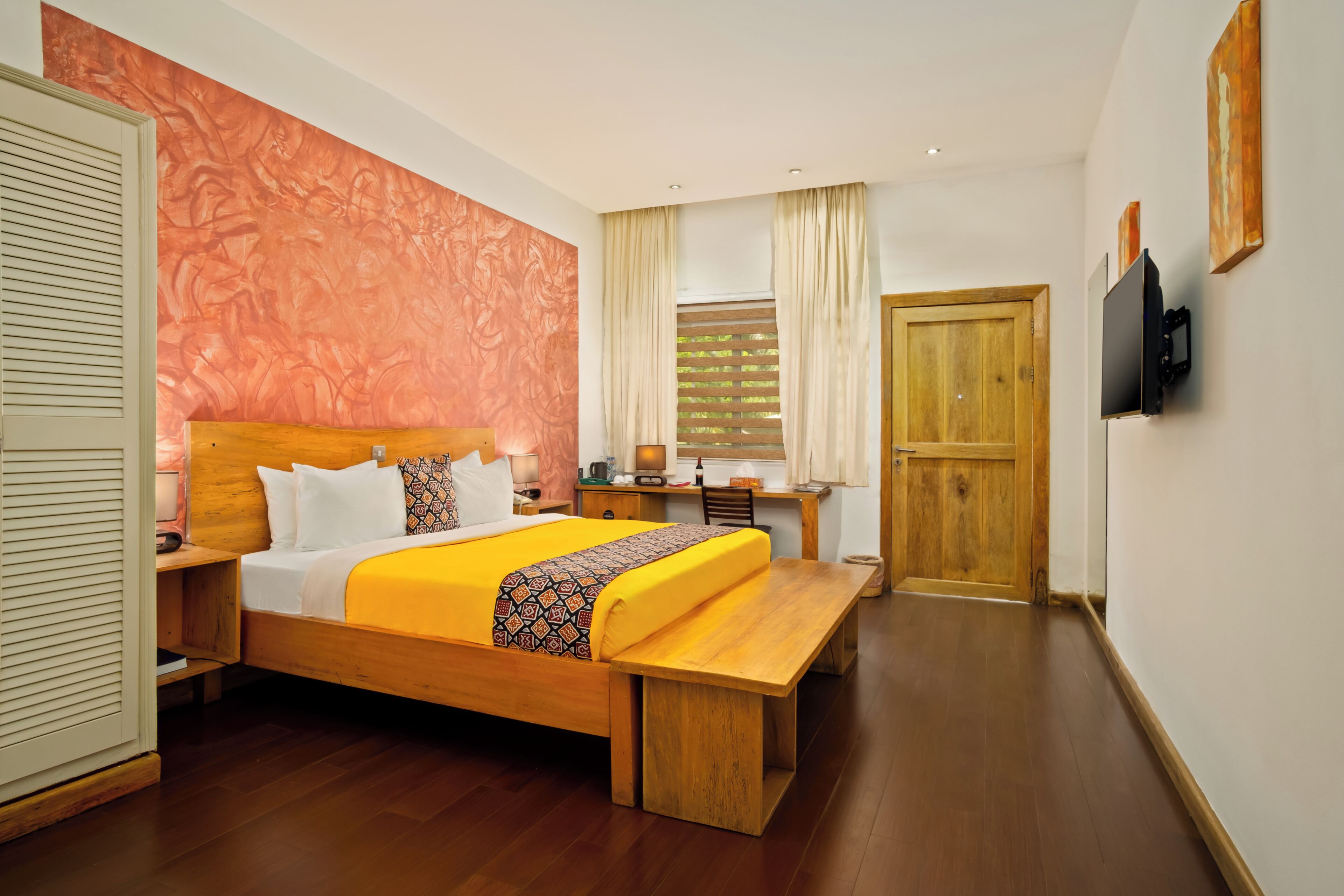 A hotel room with a wooden bed, a desk, a bench, a TV, and a door.