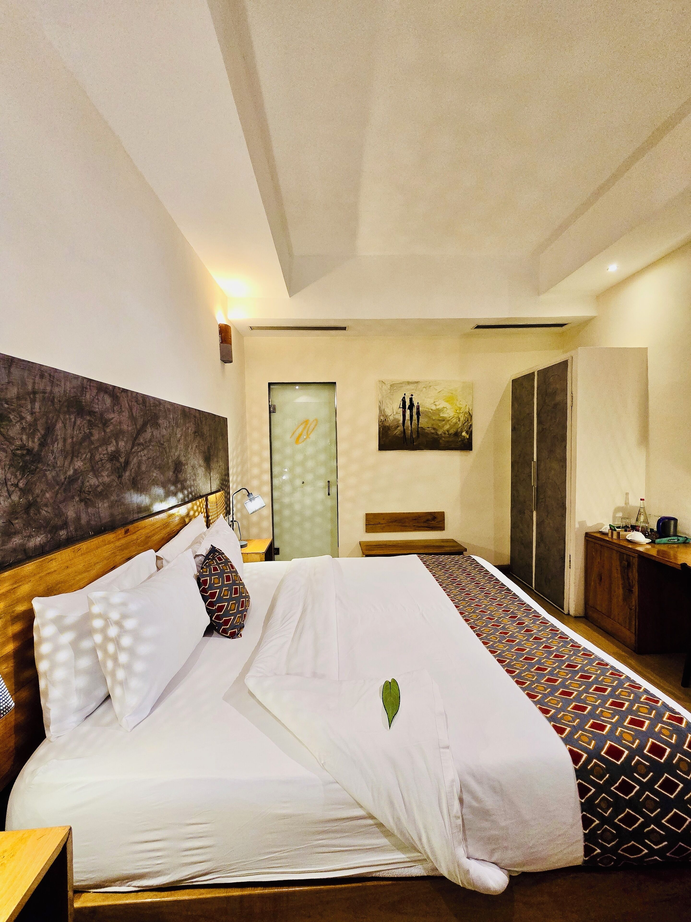 A hotel room with a large bed, a wooden headboard, a bedside table with a lamp, a wardrobe, and a painting on the wall.