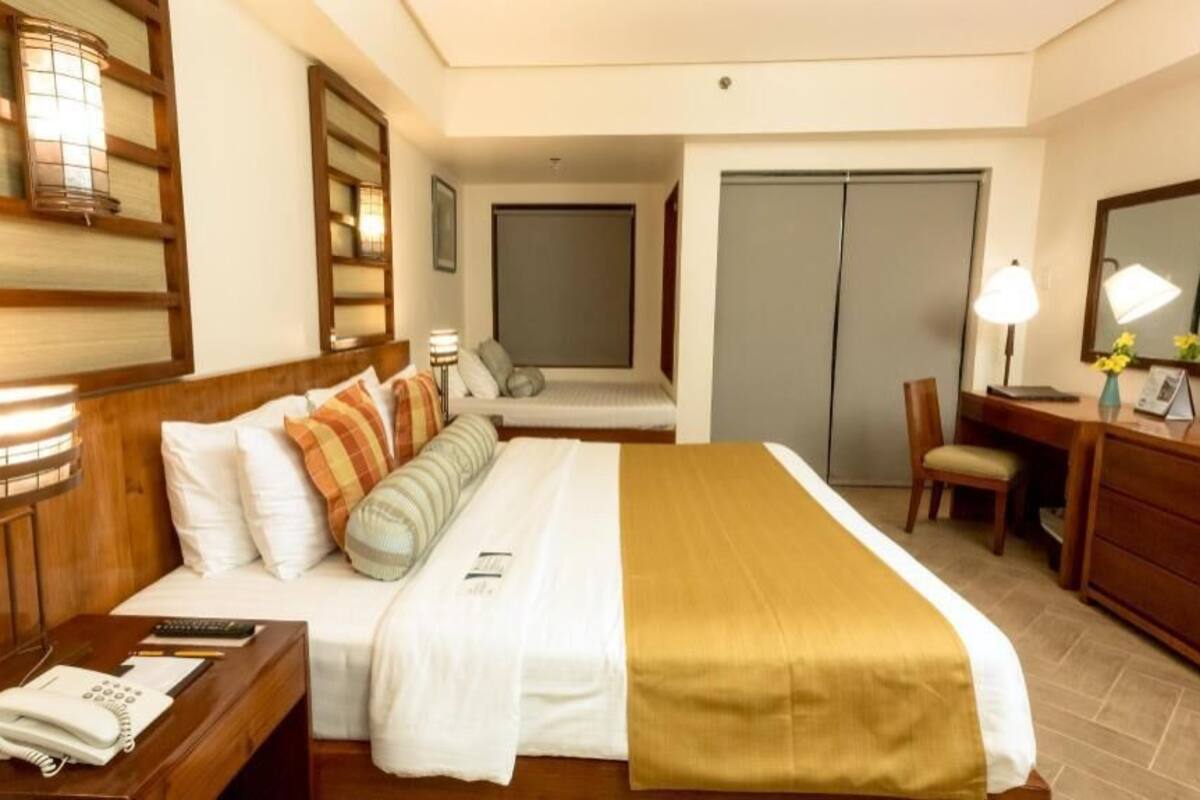 A hotel room with a bed, bedside table with a phone, a desk with a chair, a desk lamp, a mirror, and a wardrobe.