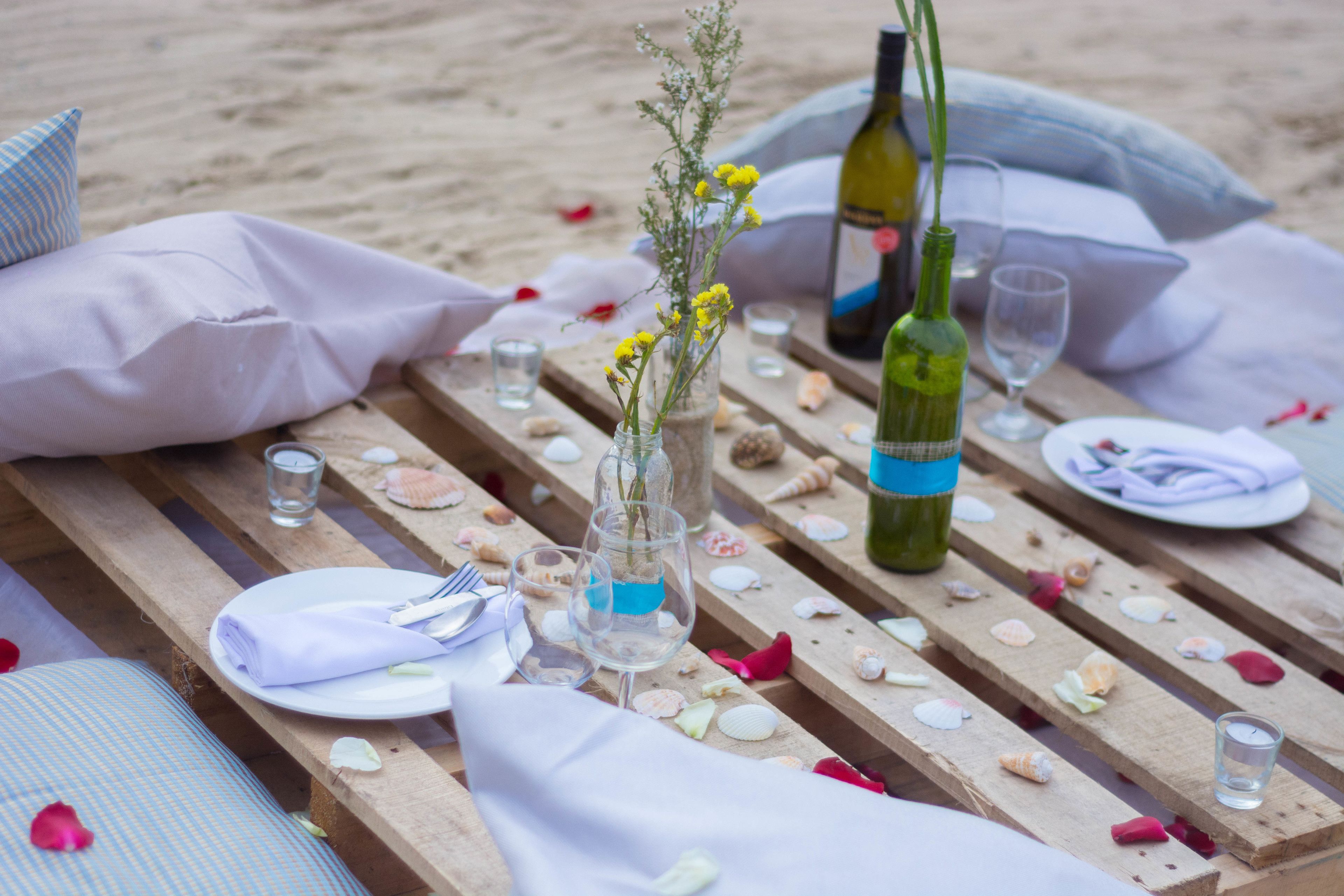 A wooden table set with a bottle, glasses, and a vase with flowers, surrounded by seashells and pillows on a beach.