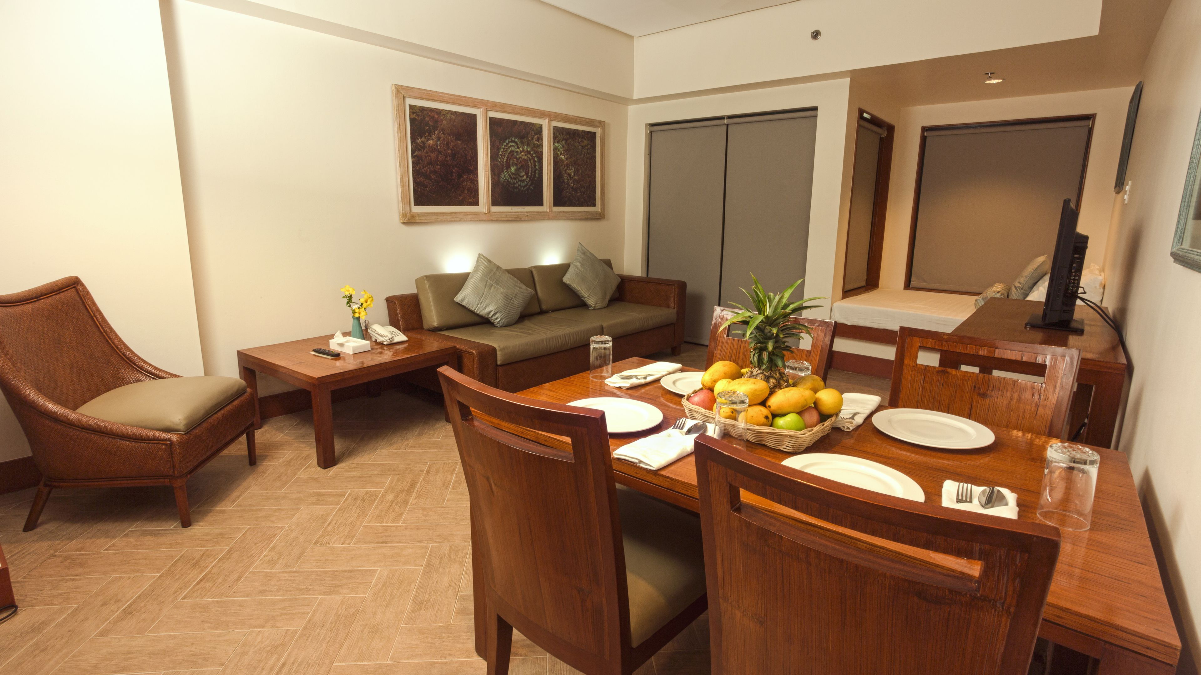 Un comedor con una mesa de madera dispuesta para cuatro personas, rodeada de sillas de madera. Hay un sofá y una mesita con un jarrón de flores y un teléfono.