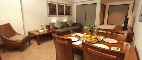 Un comedor con una mesa de madera dispuesta para cuatro personas, rodeada de sillas de madera. Hay un sofá y una mesita con un jarrón de flores y un teléfono.