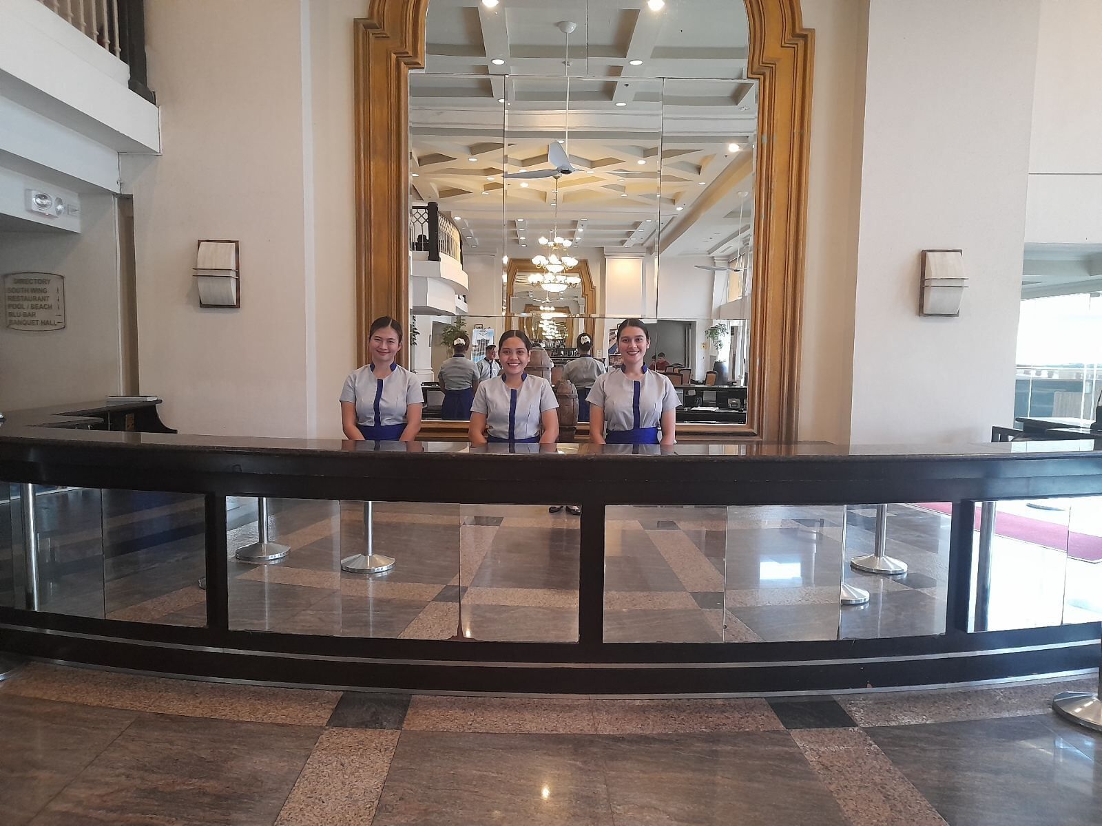 Ein Empfangsbereich mit Glastrennwand, einer Rezeption und Blick auf die Lobby mit hoher Decke und Kronleuchtern.