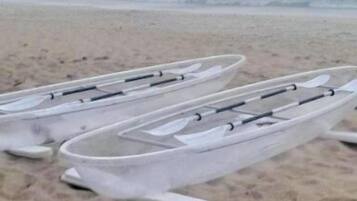 A row of white kayaks lined up on a sandy beach with a calm sea and a rocky outcrop in the background.