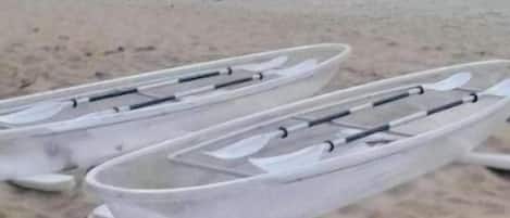 Kayaks blancos alineados en una playa arenosa, con un mar tranquilo y un promontorio rocoso al fondo.