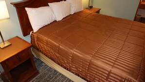 Una cama bien hecha con colcha marrón, cabecera de madera y almohadas blancas. Hay dos mesitas de noche con lámparas y un cuadro enmarcado en la pared.