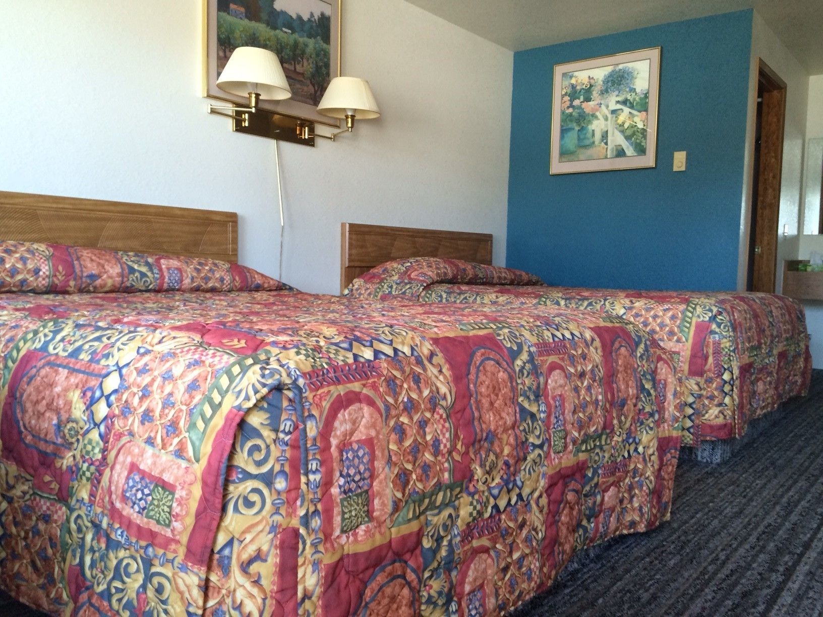 Habitación de hotel con dos camas, cada una con una colcha estampada y colorida. Hay lámparas de pared sobre las camas y un cuadro enmarcado en la pared.