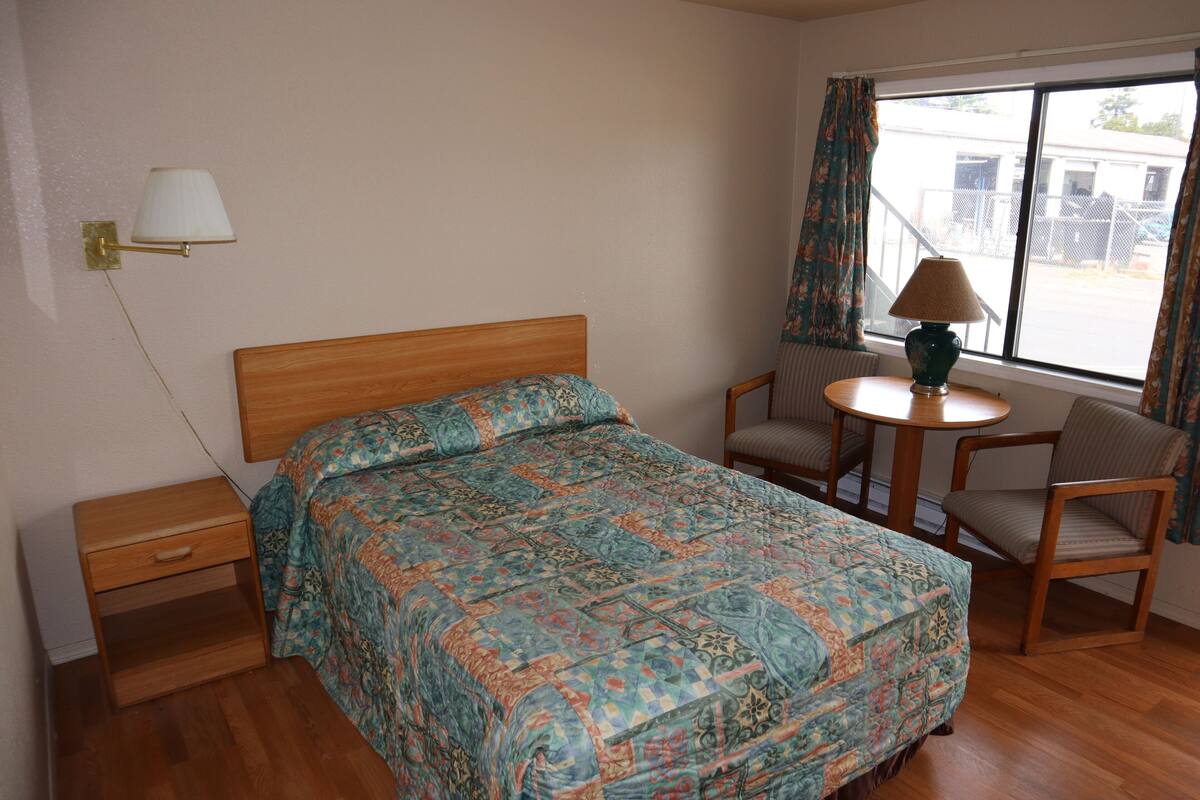 Ein ordentlich eingerichtetes Schlafzimmer mit einem hölzernen Bett, einem kleinen Nachttisch, einem Stuhl, einem runden Tisch und einem Fenster mit Vorhängen.