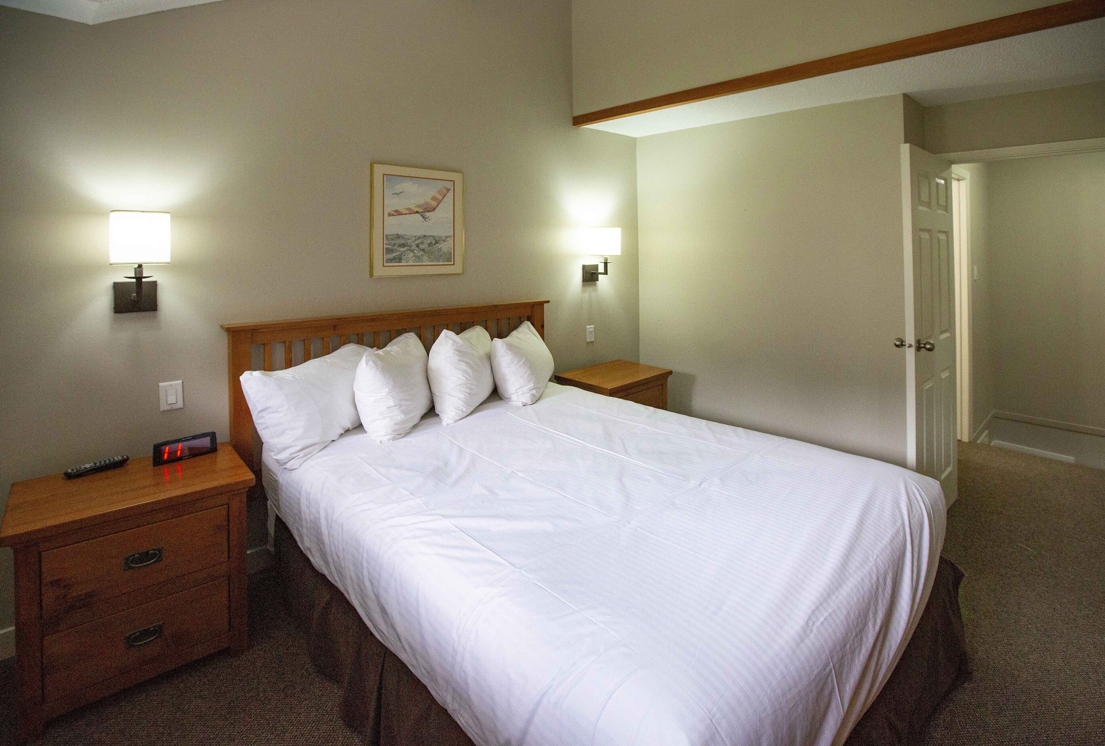 A neatly made bed with white linens, two bedside tables with lamps, a framed picture on the wall, and a door to the right.