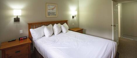 A neatly made bed with white linens, two bedside tables with lamps, a framed picture on the wall, and a door to the right.