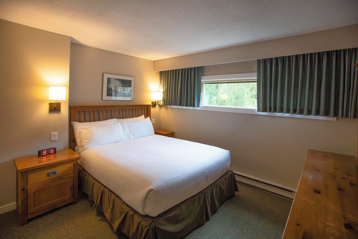 Habitación de hotel con una cama grande, dos mesitas de noche con lámparas, una ventana con cortinas y un reloj en la pared.