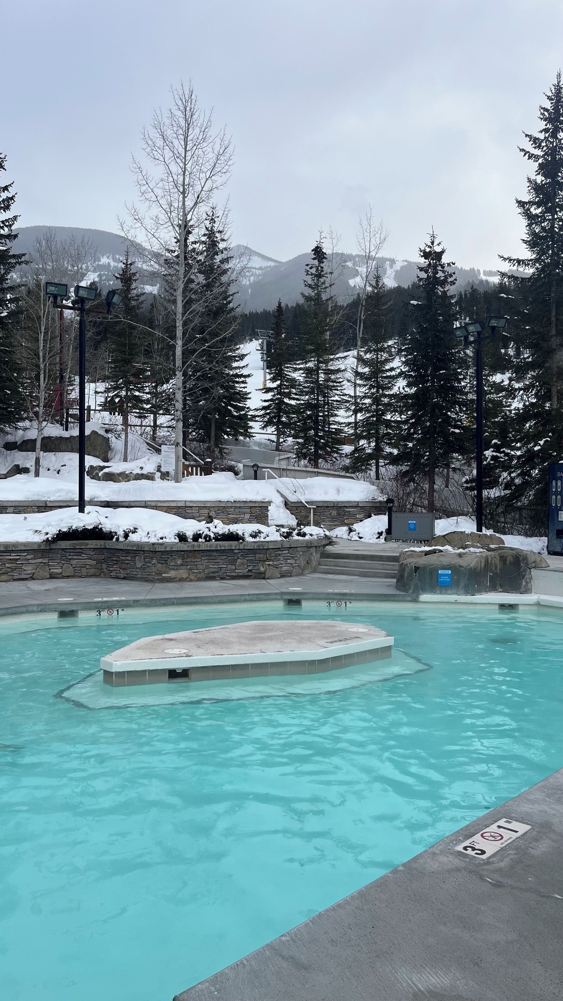 A swimming pool with clear water, surrounded by snow-covered ground and trees, with mountains in the background.