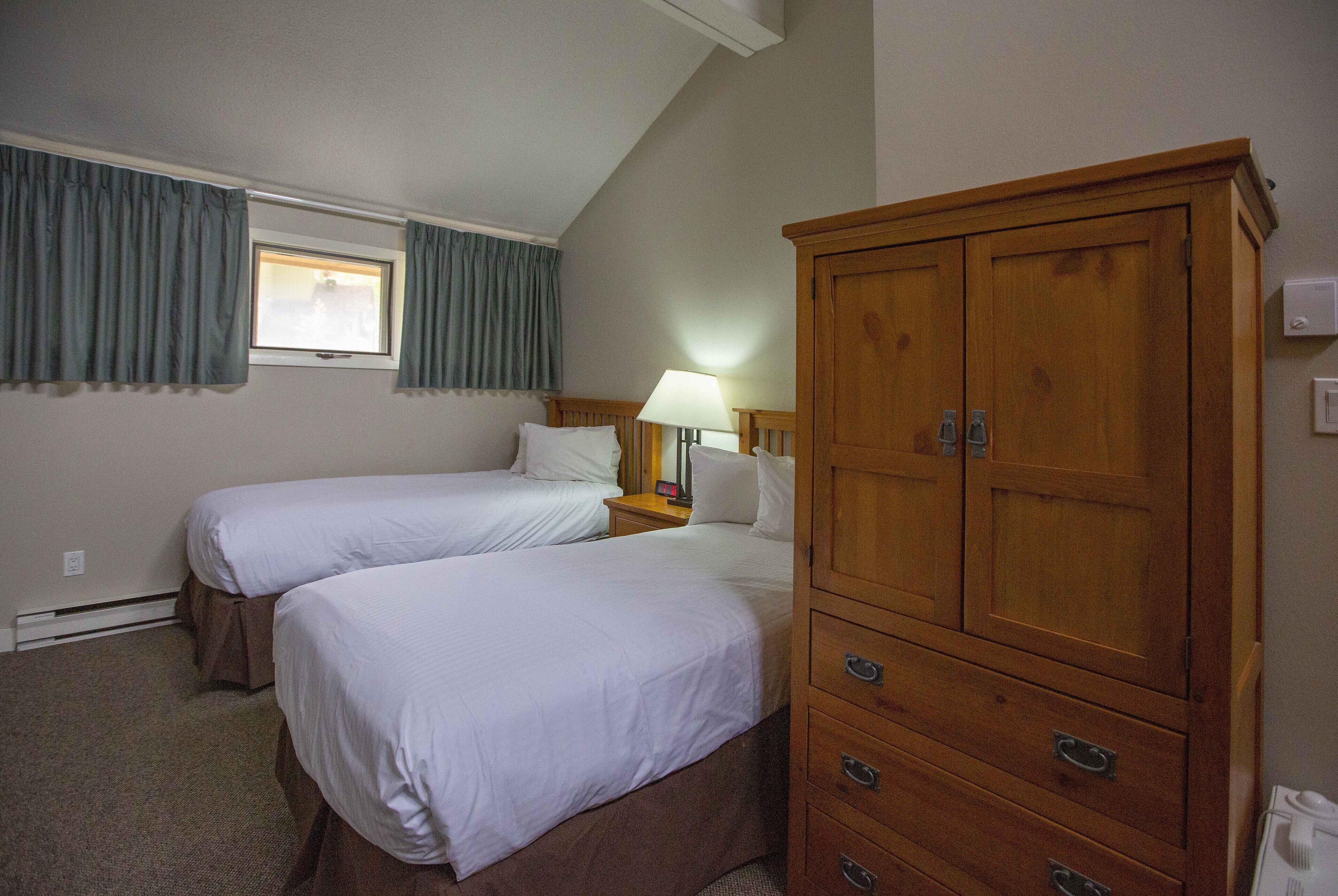 A hotel room with two beds, a wooden wardrobe, a nightstand with a lamp, and a window with curtains.