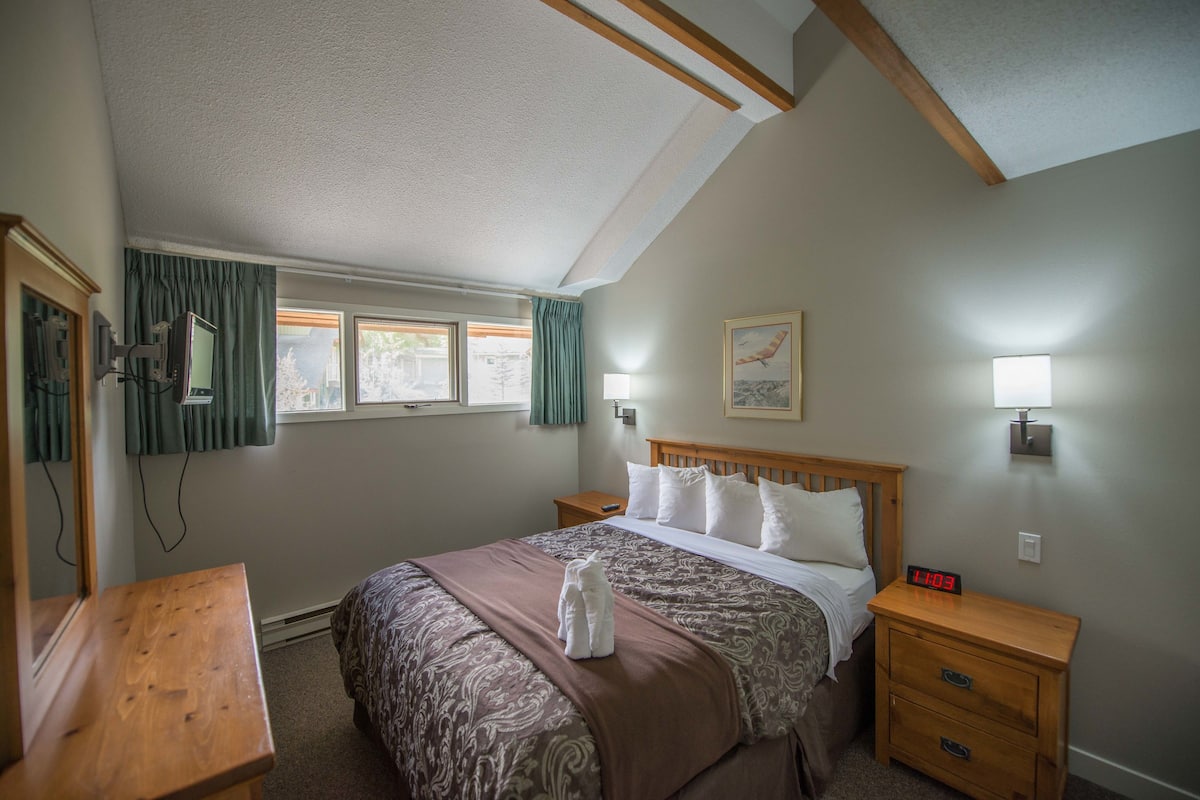 Una habitación de hotel con cama, mesitas de noche, una mesita con un reloj, lámpara, ventana con cortinas y un cuadro en la pared.