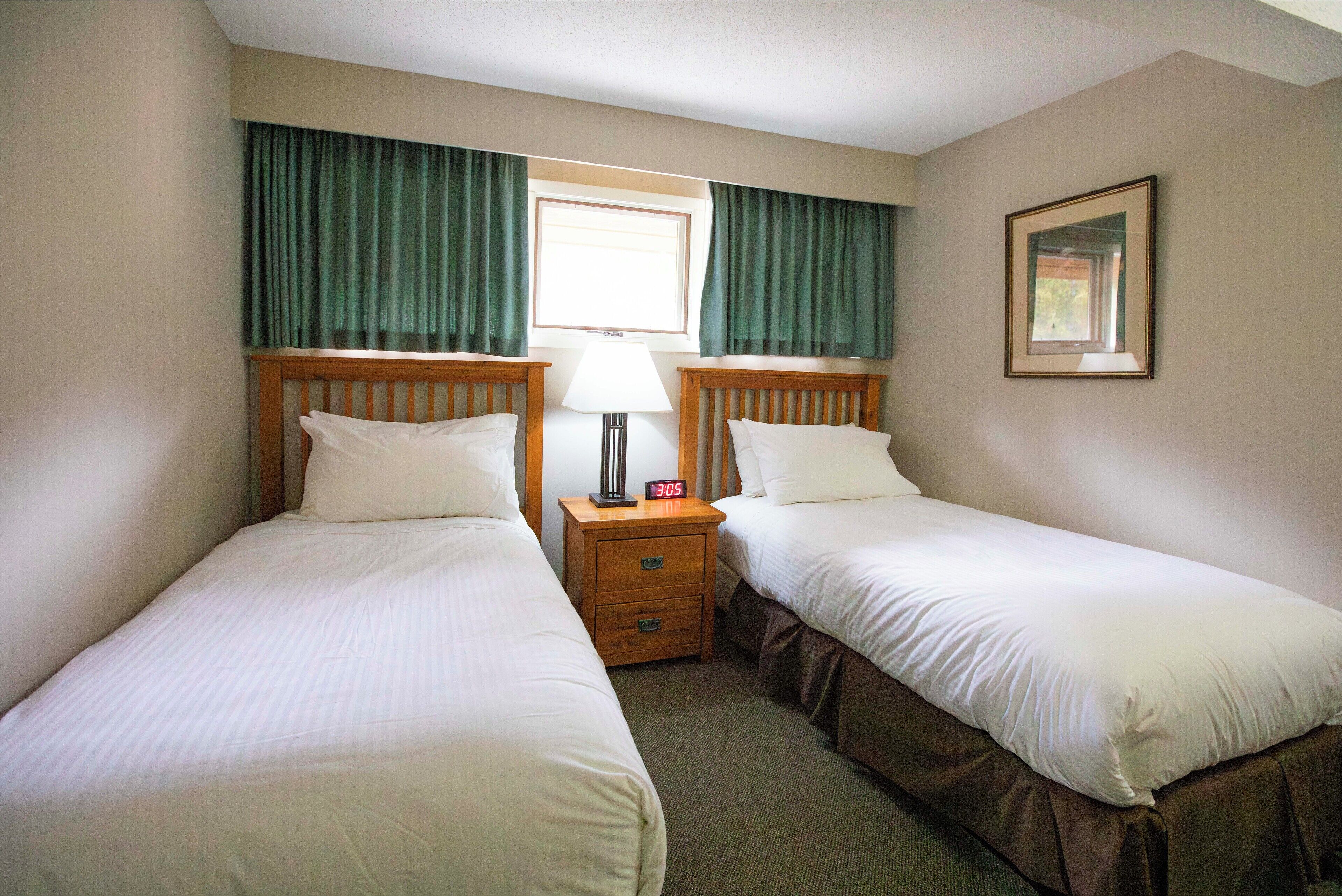 Quarto de hotel com duas camas de solteiro, mesa de cabeceira com abajur, relógio e janela com cortinas.