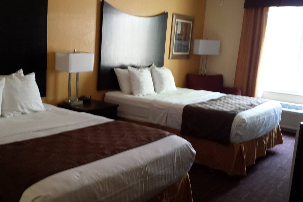 A hotel room with two beds, a nightstand, a lamp, and a window with curtains.