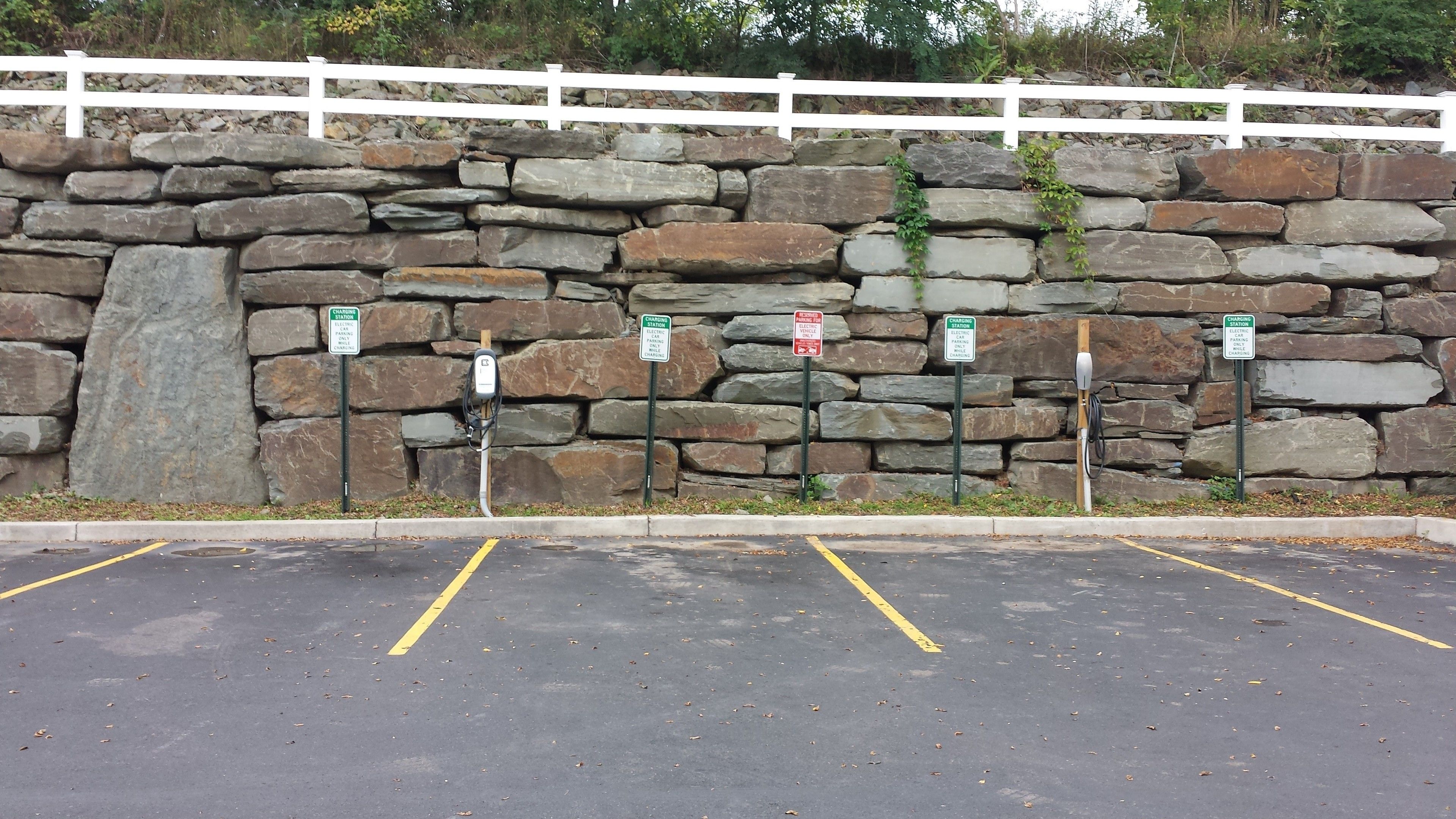 A parking area with electric vehicle charging stations, a stone wall, and a white fence.