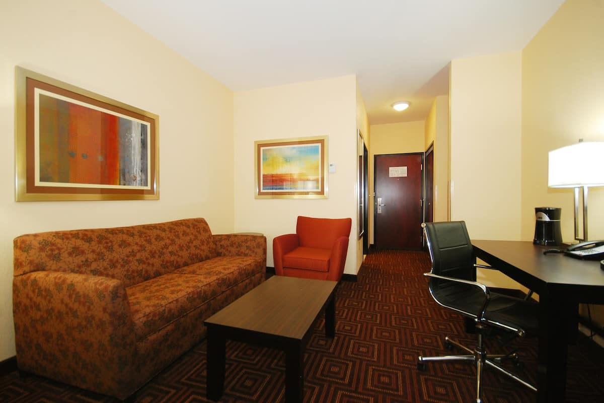 A hotel room with a floral sofa, a small red armchair, a wooden coffee table, a black office chair, a desk with a lamp, and two framed artworks on the walls.