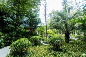 Un jardin luxuriant, avec différents arbres et arbustes, un sentier et un bâtiment en arrière-plan.