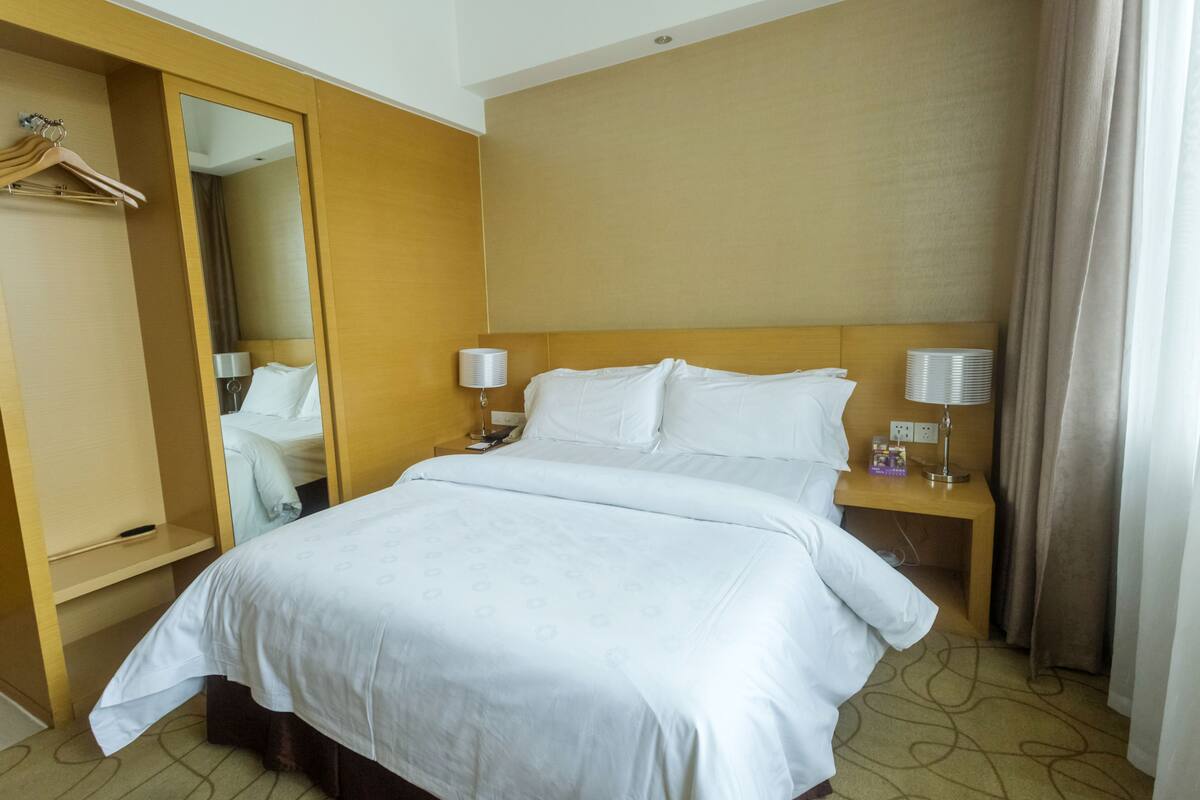 Une chambre d’hôtel avec un grand lit, deux tables de chevet, un miroir, un porte-manteau et une fenêtre avec des rideaux.