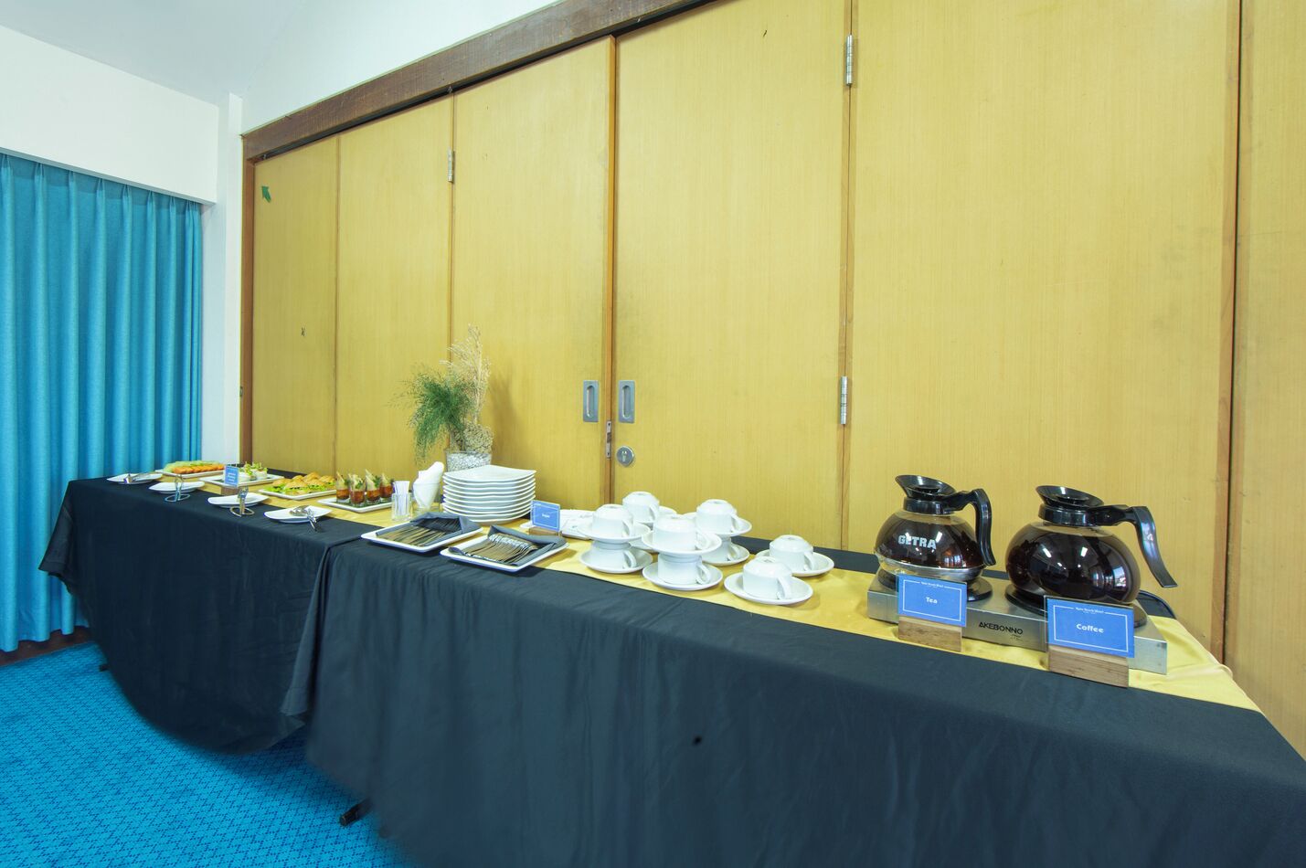 A conference room with a long table set for a meeting, equipped with coffee and tea stations, and a large wooden wardrobe in the background.