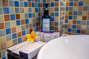 A bathroom with a tiled wall, a bottle of hand soap, a yellow flower, and boxes of toiletries. - Kuta Beach Hotel (Kuta)
