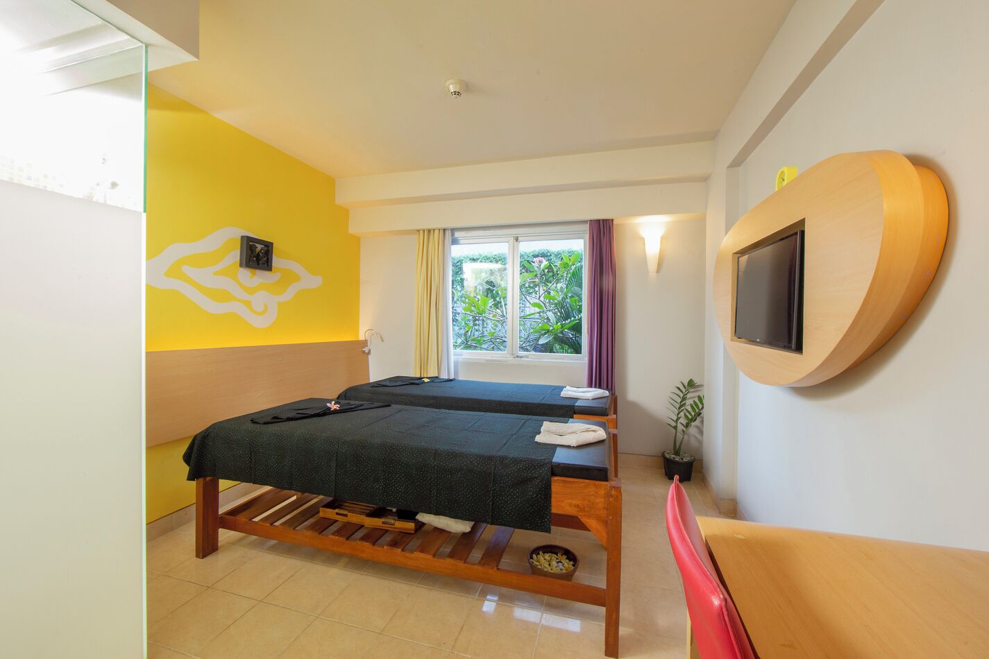 A room with two beds, a TV mounted on the wall, a window with curtains, and a potted plant.