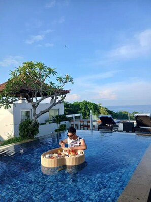 A person dining at a poolside table with a wicker basket containing food, surrounded by lounge chairs and a view of the sea.