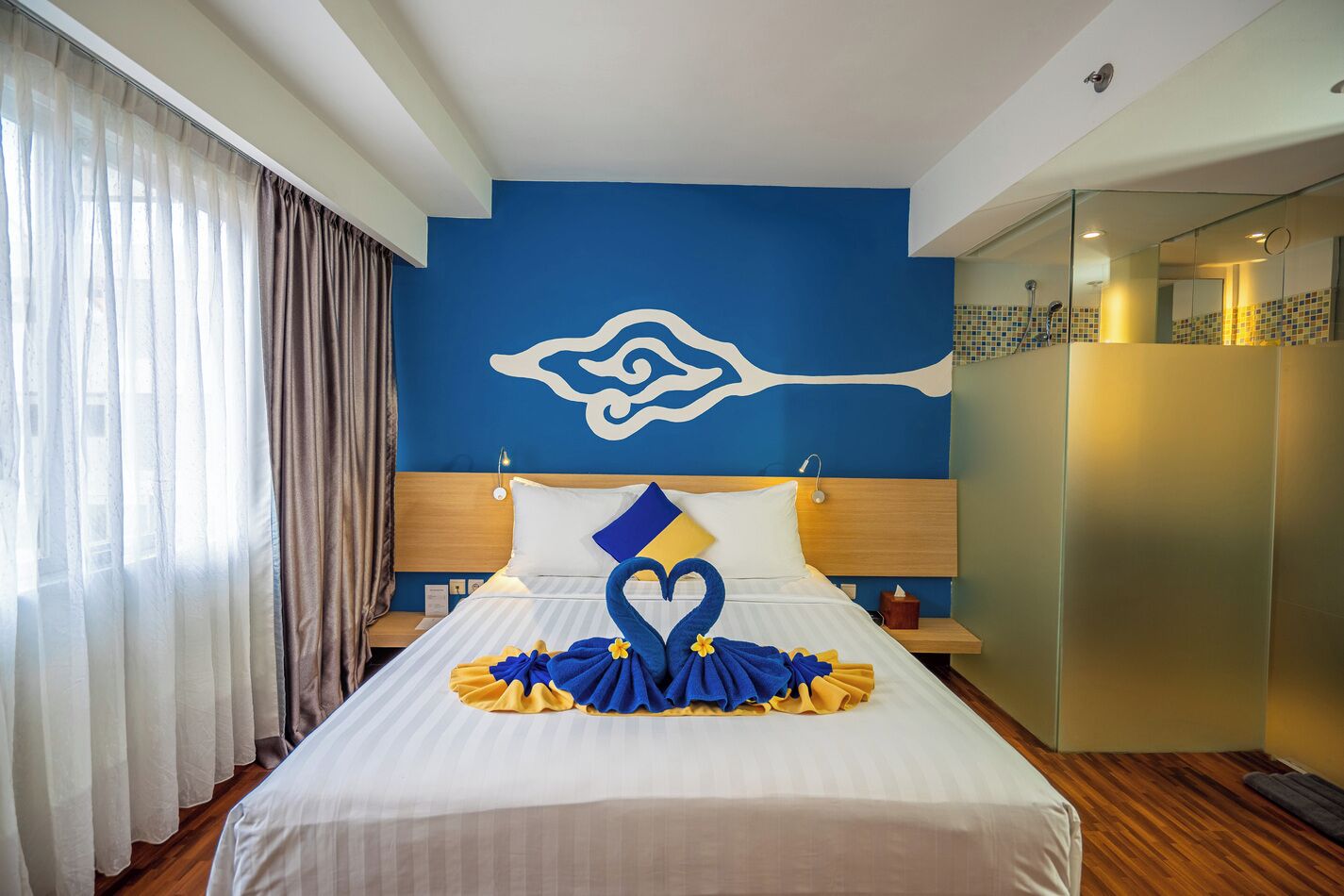 Une chambre d’hôtel avec un grand lit, une tête de lit ornée d’un motif décoratif, une table de chevet et une salle de bain visible à travers une porte en verre.