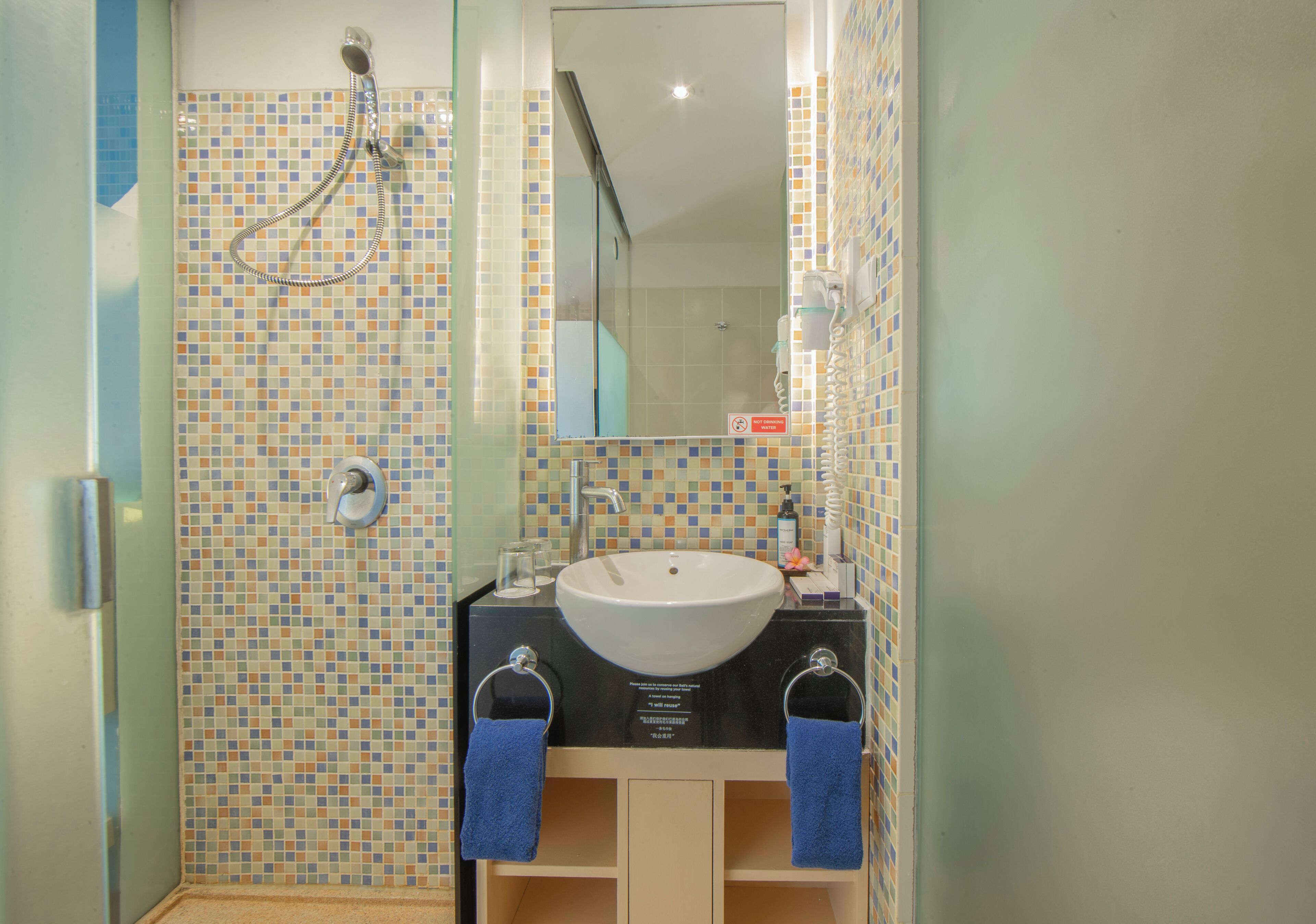 A bathroom with a tiled shower wall, a sink with a mirror, and two towels hanging on hooks.