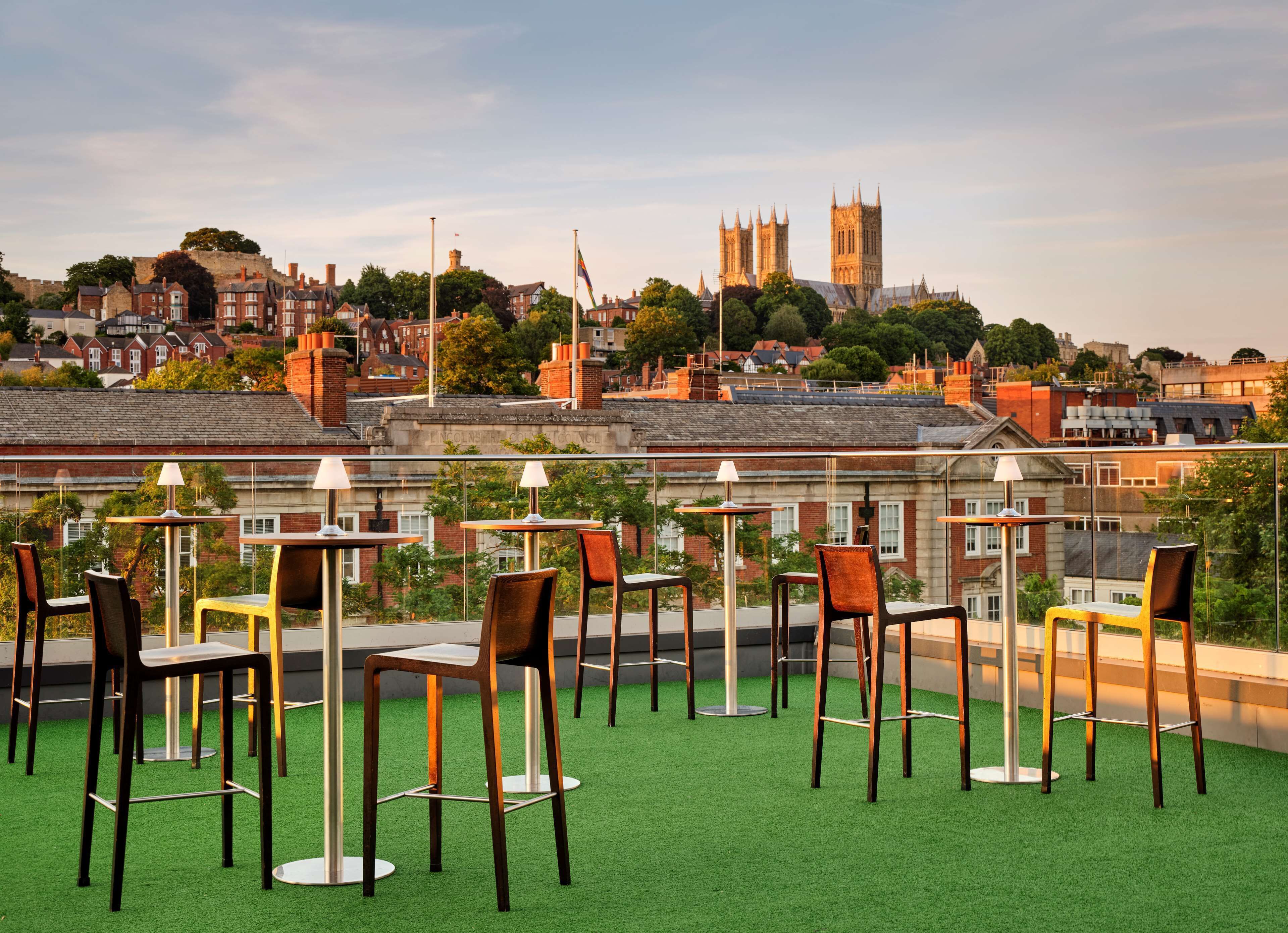 Une terrasse sur le toit, aménagée avec des chaises et des tables, offrant une vue sur un paysage urbain historique, dominé par le clocher d’une église remarquable.