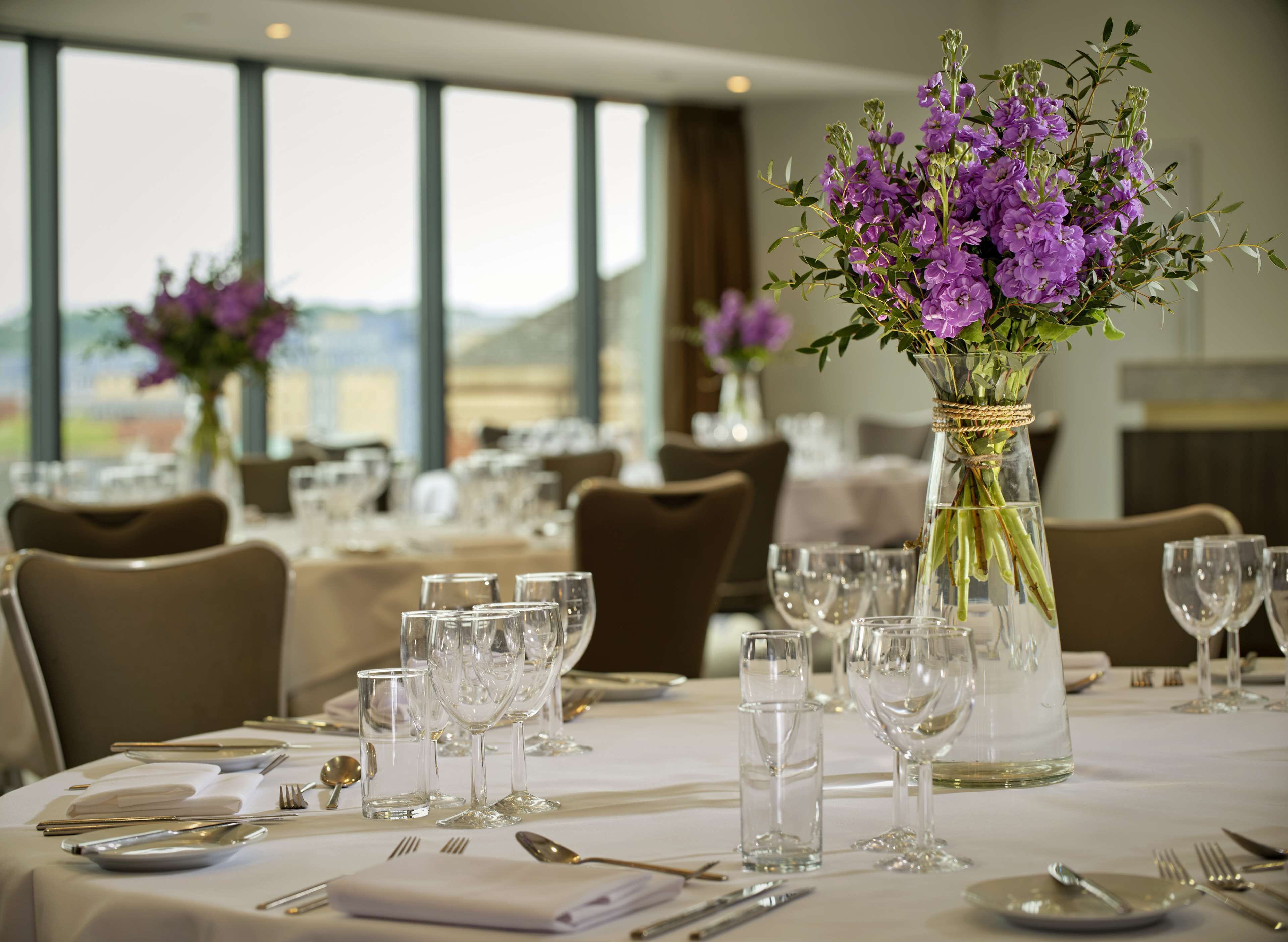 A dining area with tables set for a meal, featuring clear glassware and purple flowers in vases.
