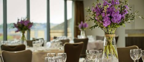A dining area with tables set for a meal, featuring clear glassware and purple flowers in vases.