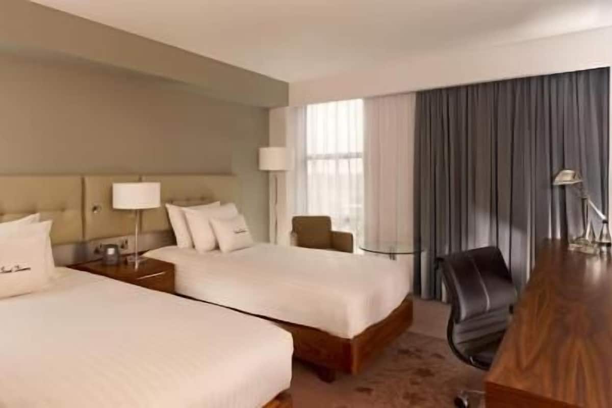 Habitación de hotel con dos camas, un escritorio, una silla y un ventanal con cortinas.