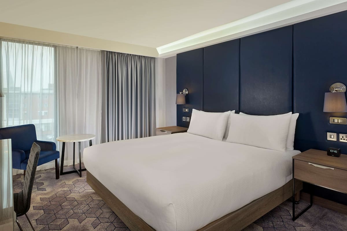 A hotel room with a large bed, a desk with a lamp, a chair, and a small table.