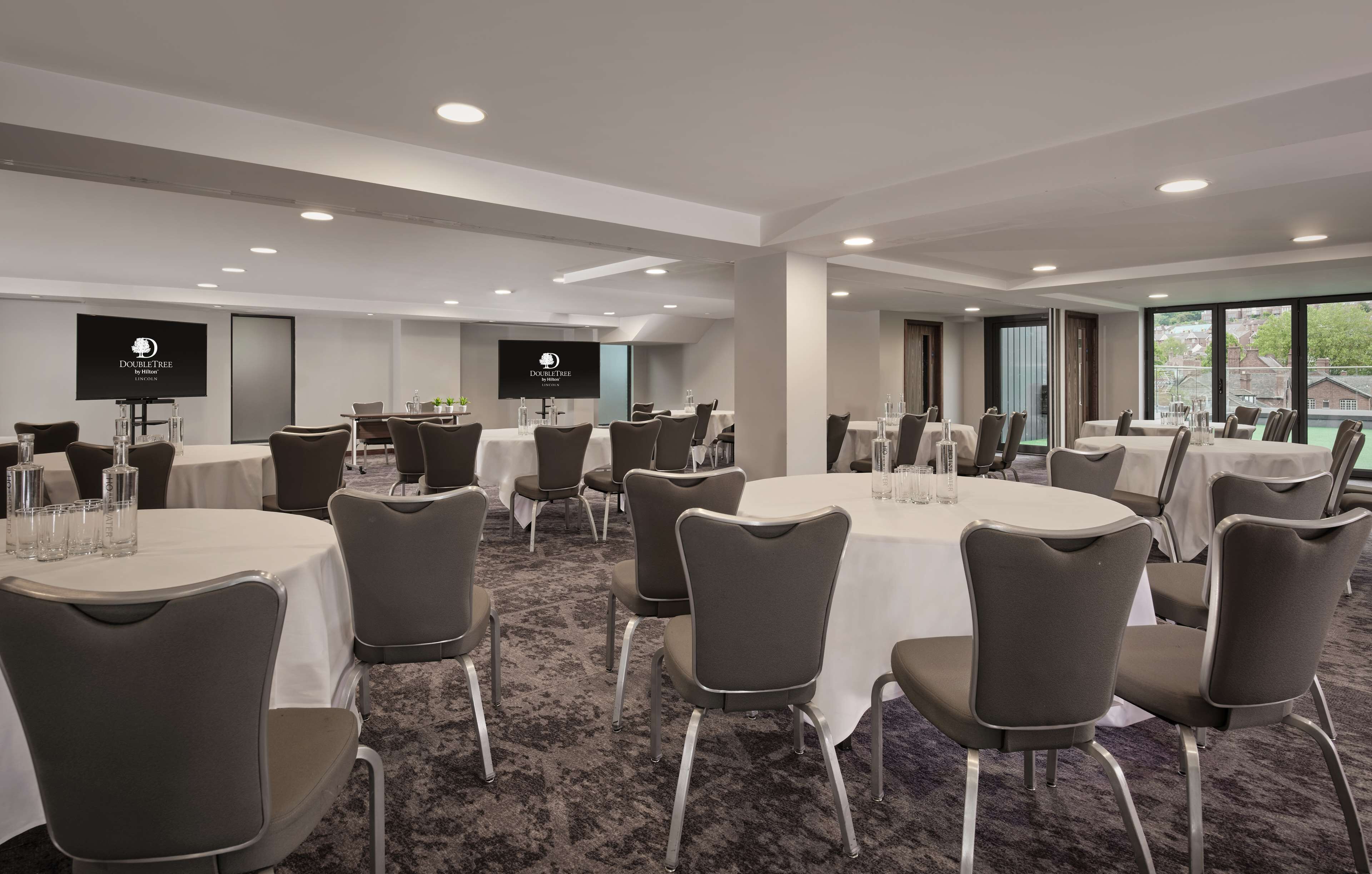 Ein Konferenzraum mit runden Tischen, grauen Stühlen und einer großen Leinwand, auf der das DoubleTree-Logo zu sehen ist.