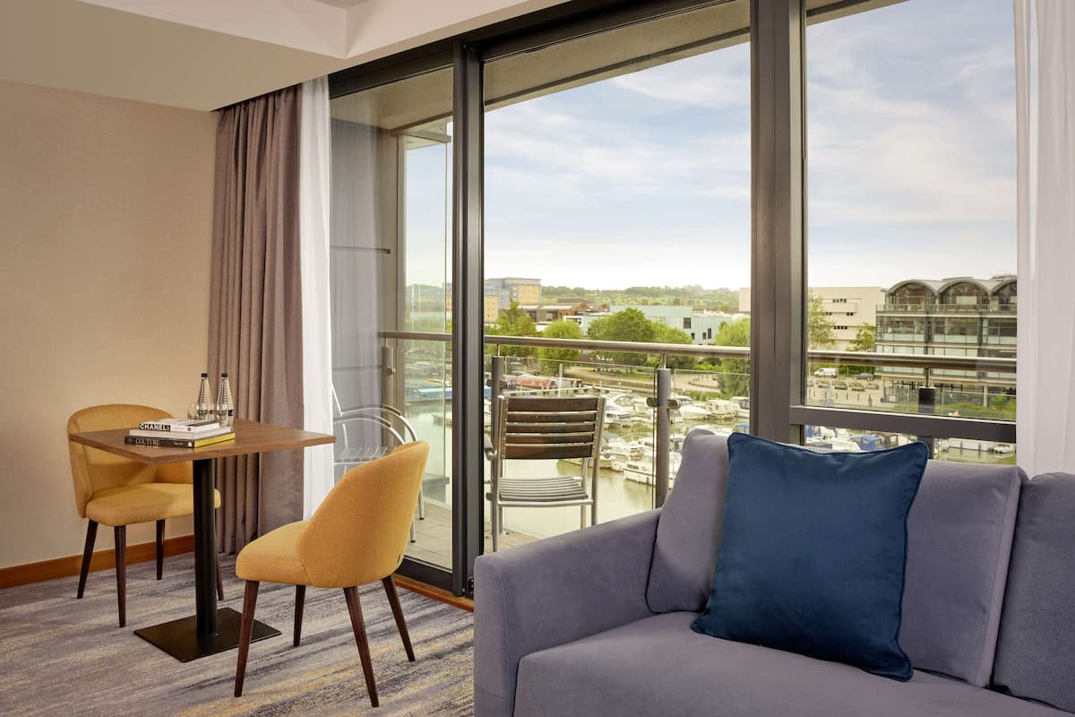 A modern hotel room with a balcony, a sofa, a dining table with chairs, and a view of the cityscape.