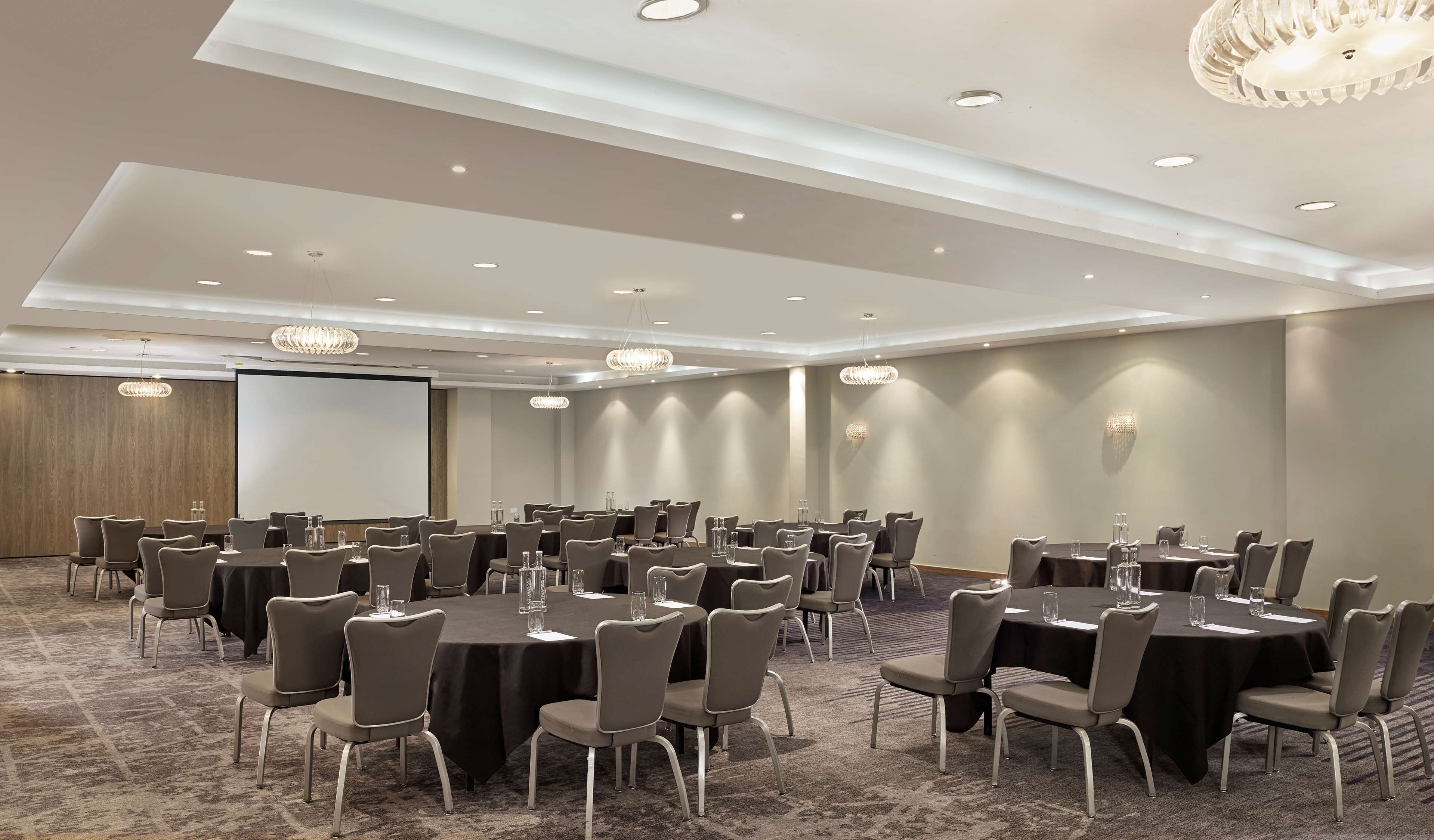 A conference room with multiple tables and chairs, a projector screen, and a carpeted floor.