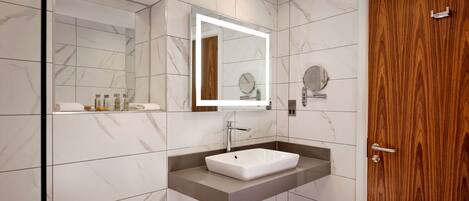 Un baño moderno con pared de azulejos de mármol, puerta de madera, lavamanos blanco con espejo y un perchero.